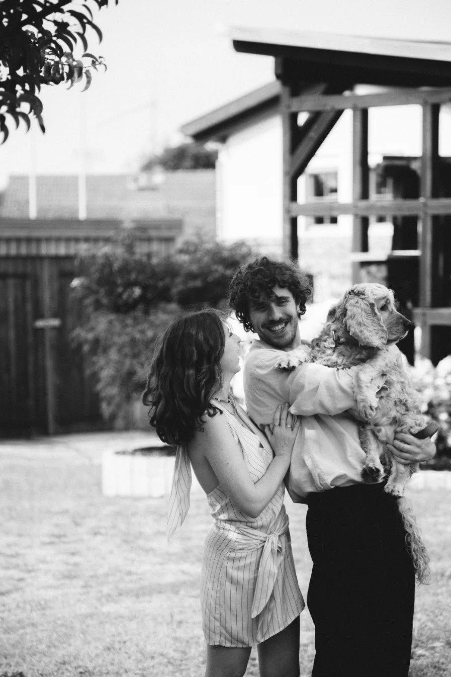 A medium black and white outdoor portrait of a smiling woman and a man holding a fluffy dog while looking at each other.