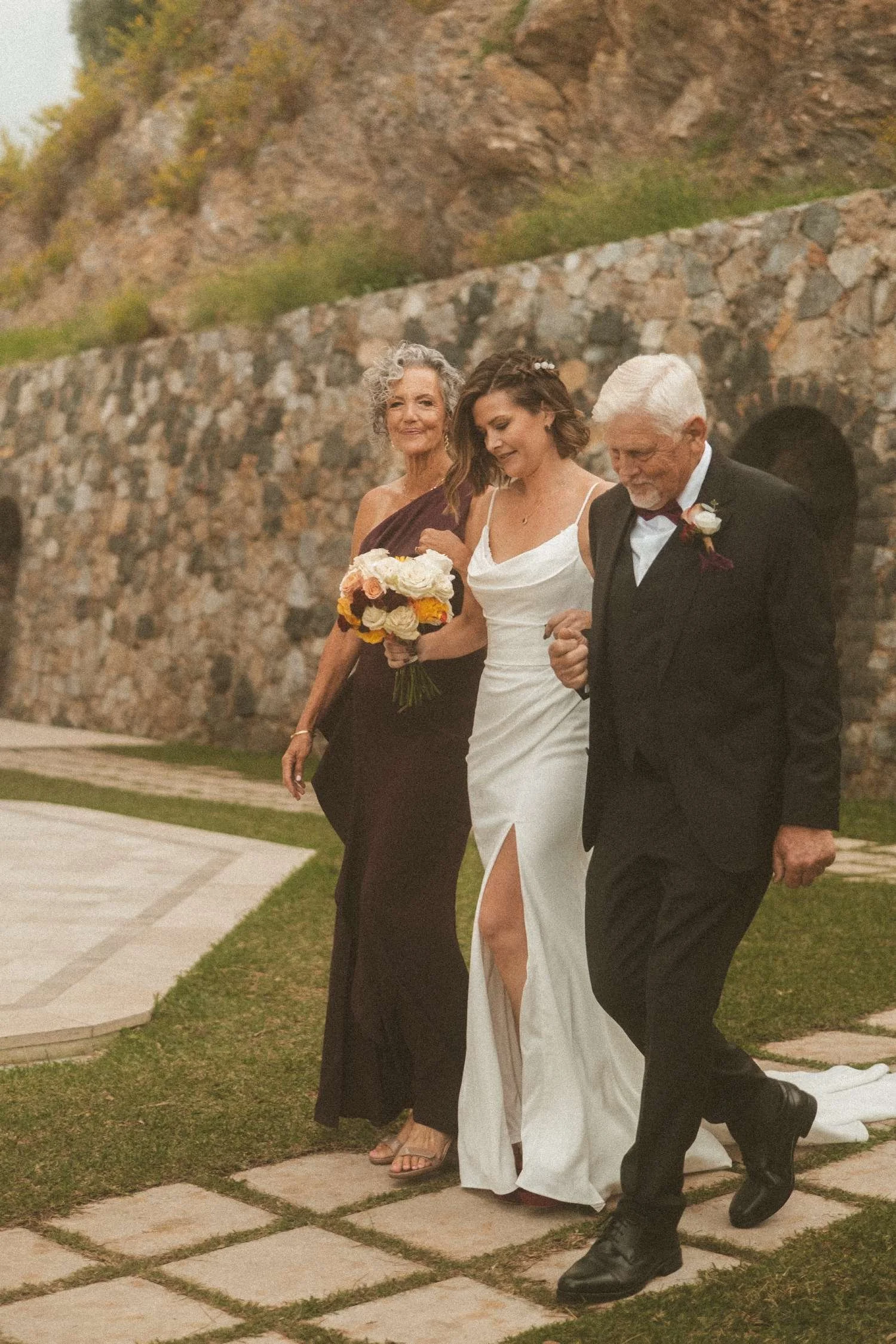 Bride walking arm in arm with her parents during an intimate outdoor wedding ceremony beside a stone wall.