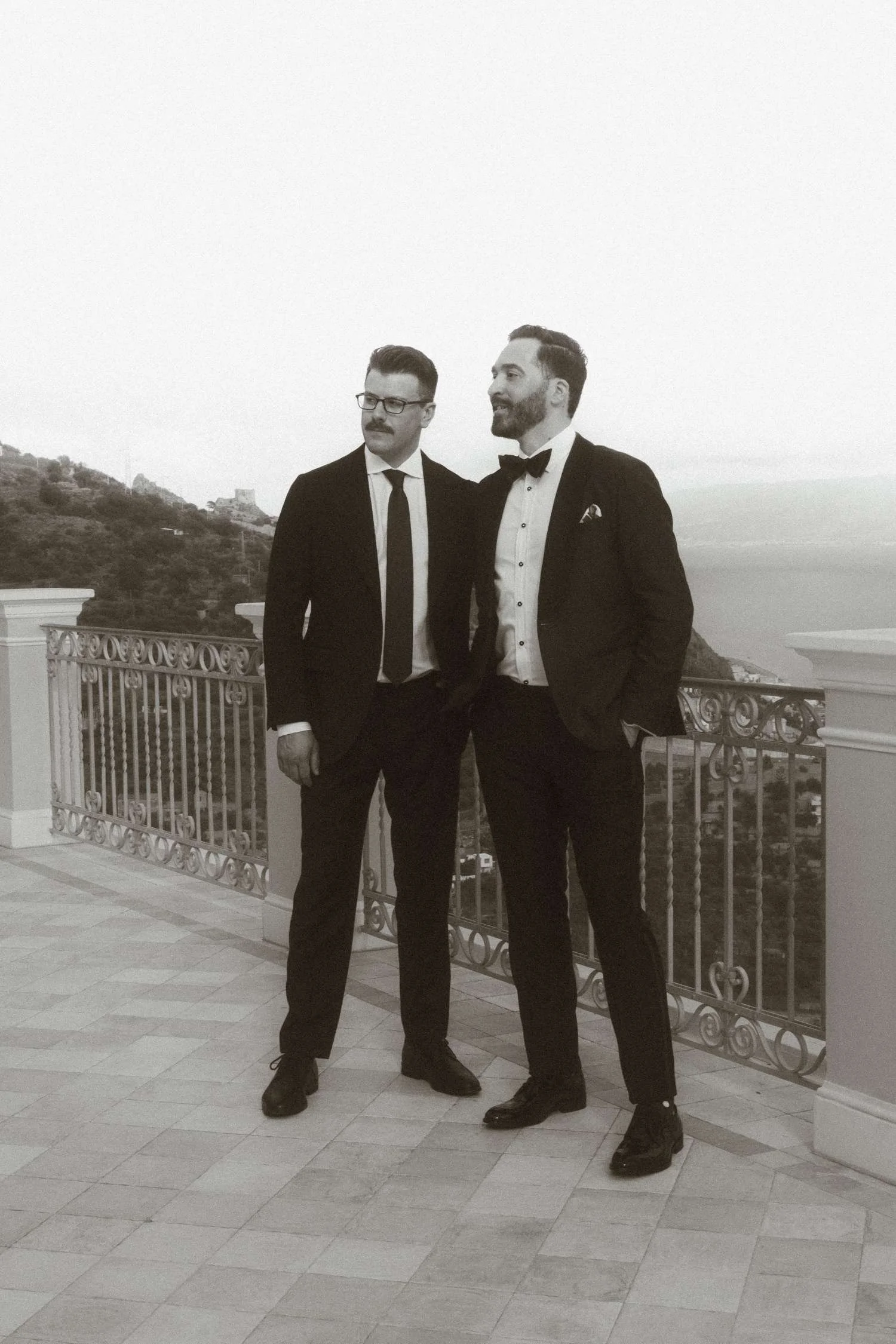 Two grooms in formal black suits standing together on a scenic Italian wedding terrace, captured in an elegant black and white portrait.