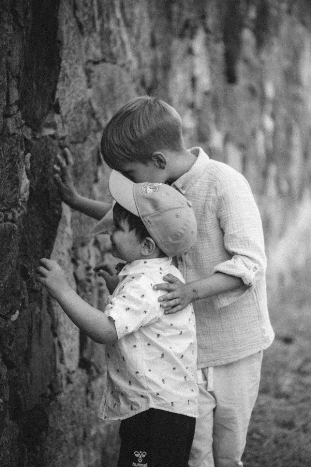 A medium black and white outdoor shot of two young boys playing and touching a rustic stone wall together.
