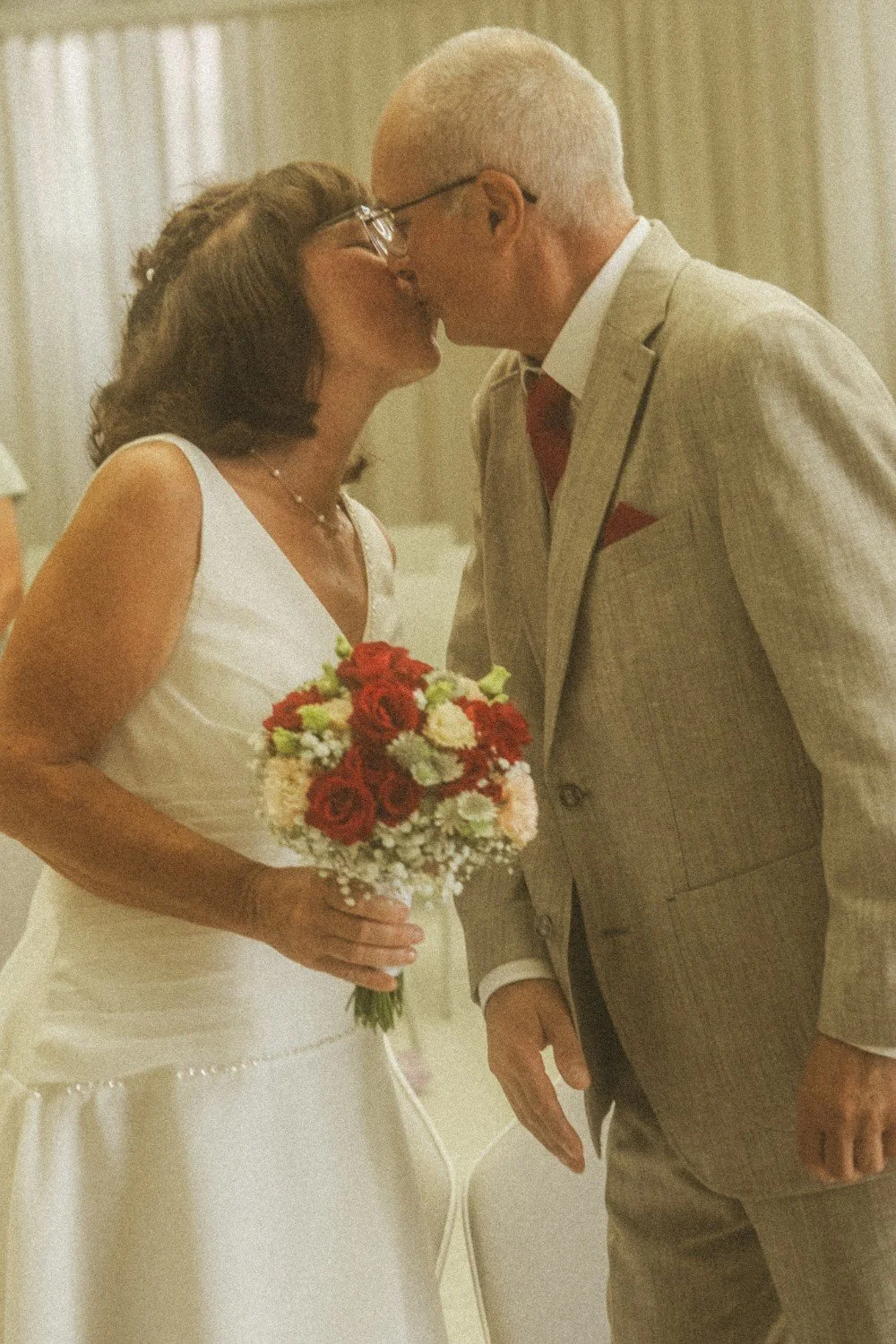 A close-up indoor shot of the wedding couple sharing a kiss, with the bride holding a bouquet of red and white flowers.