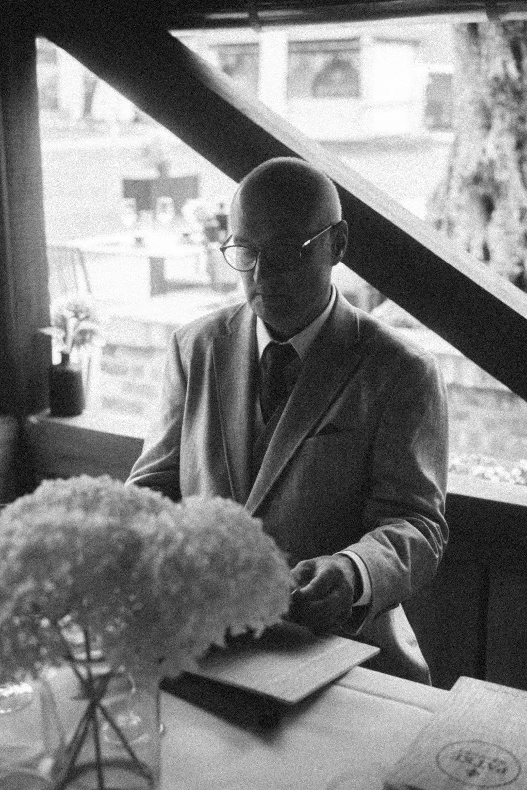 A black and white portrait of a groom in a suit looking down at a menu at a table.