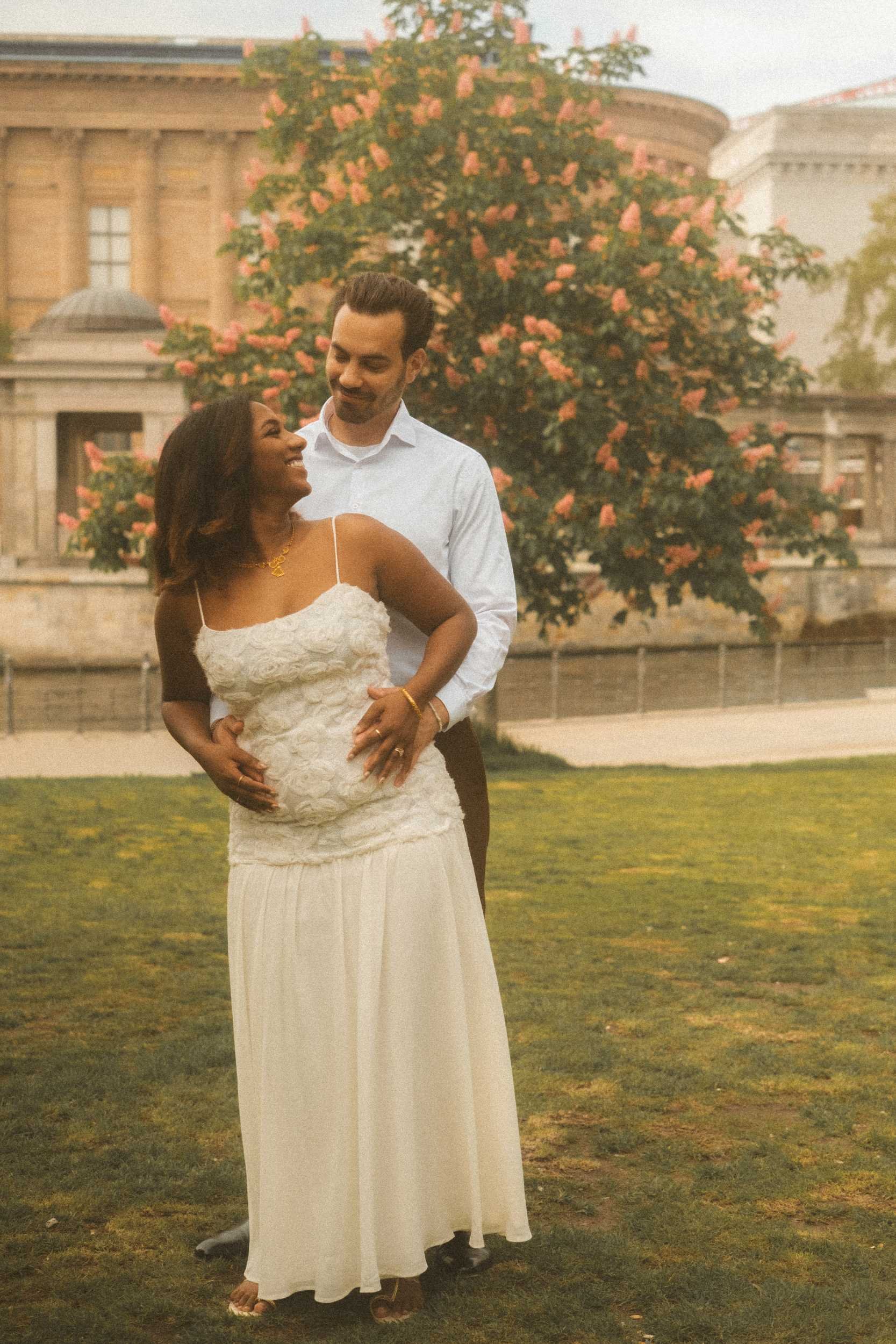A romantic engagement portrait of a couple in Berlin, with the groom embracing the bride from behind while they share an intimate moment of eye contact.