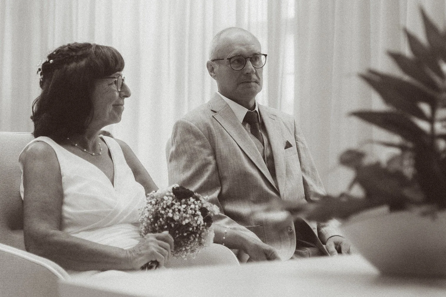 A vintage-style black and white portrait of a bride holding a bouquet and a groom seated indoors together.