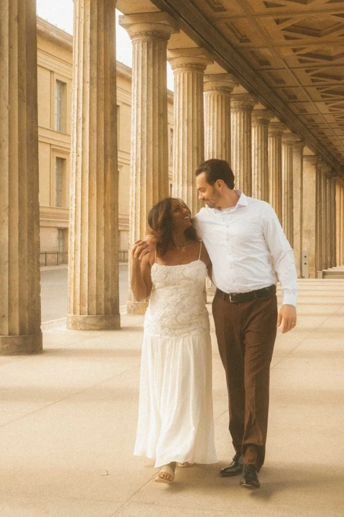 A happy couple walking arm in arm through a historic neoclassical colonnade during a golden hour engagement photoshoot in Berlin.