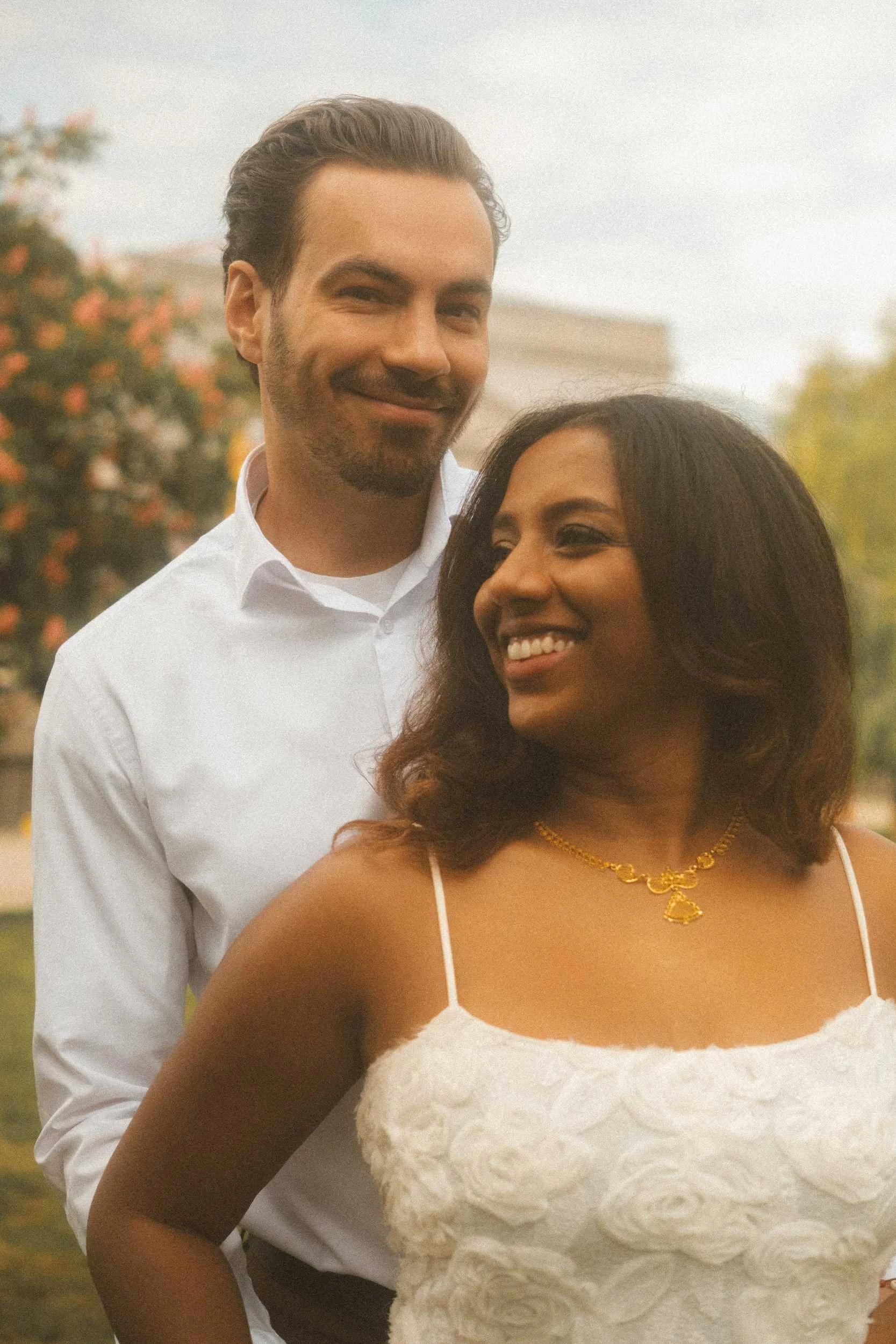 An engagement portrait in Berlin featuring the groom embracing the bride from behind while looking into the camera with a smile, as she gazes to the side with a smile.