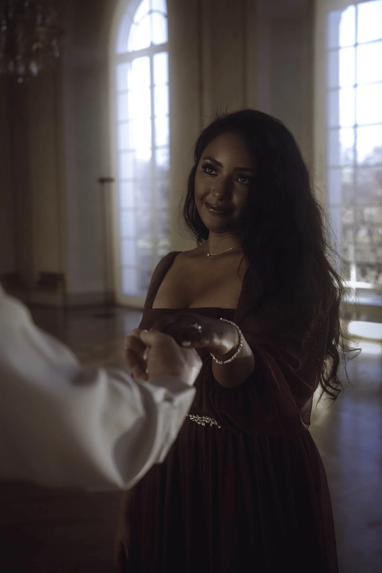 An intimate and romantic engagement photo capturing a tender moment between the couple inside the stunning Charlottenburg Palace in Berlin. The bride-to-be is elegantly holding her partner's hand, with soft natural light streaming through the grand w