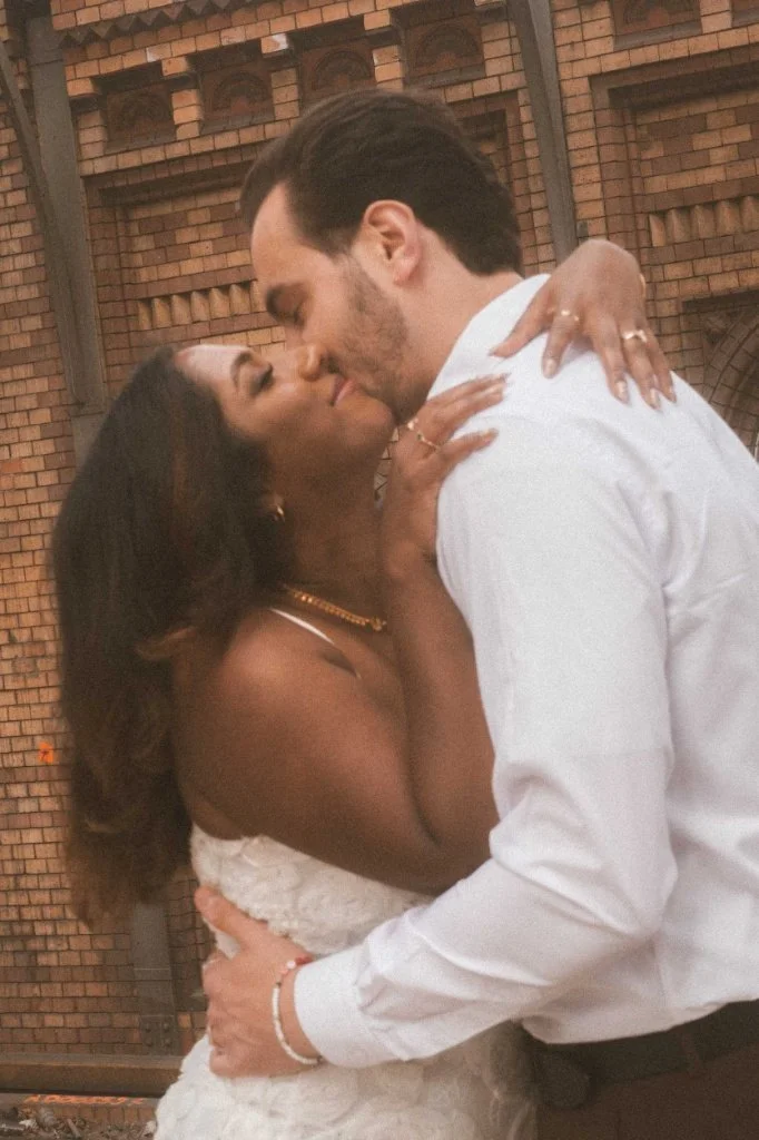 A romantic close-up of a couple sharing a tender kiss during an engagement photoshoot set against an urban brick backdrop in Berlin.