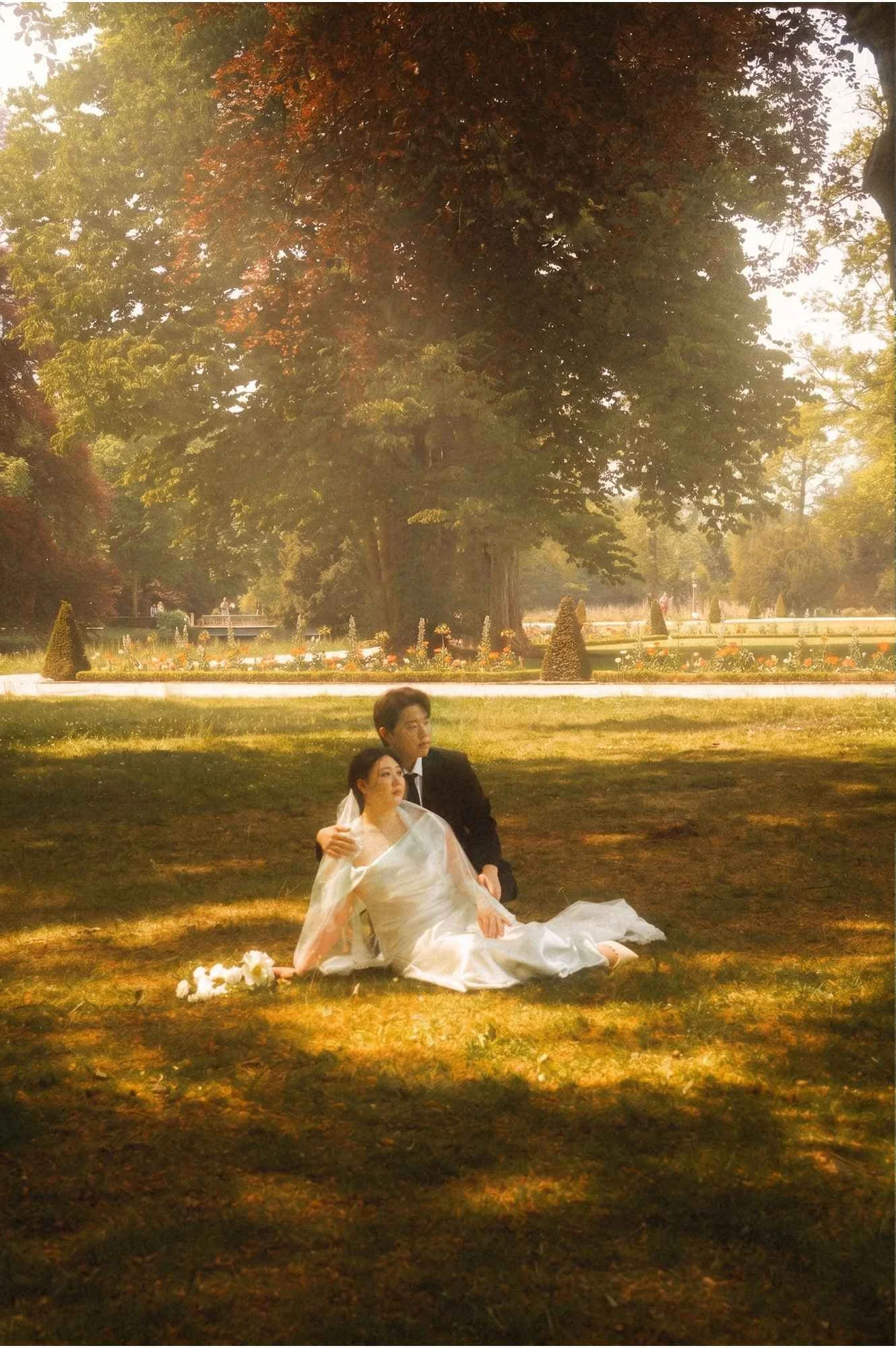 A bride in a white dress and a groom in a dark suit sitting together on the grass outdoors in Potsdam.