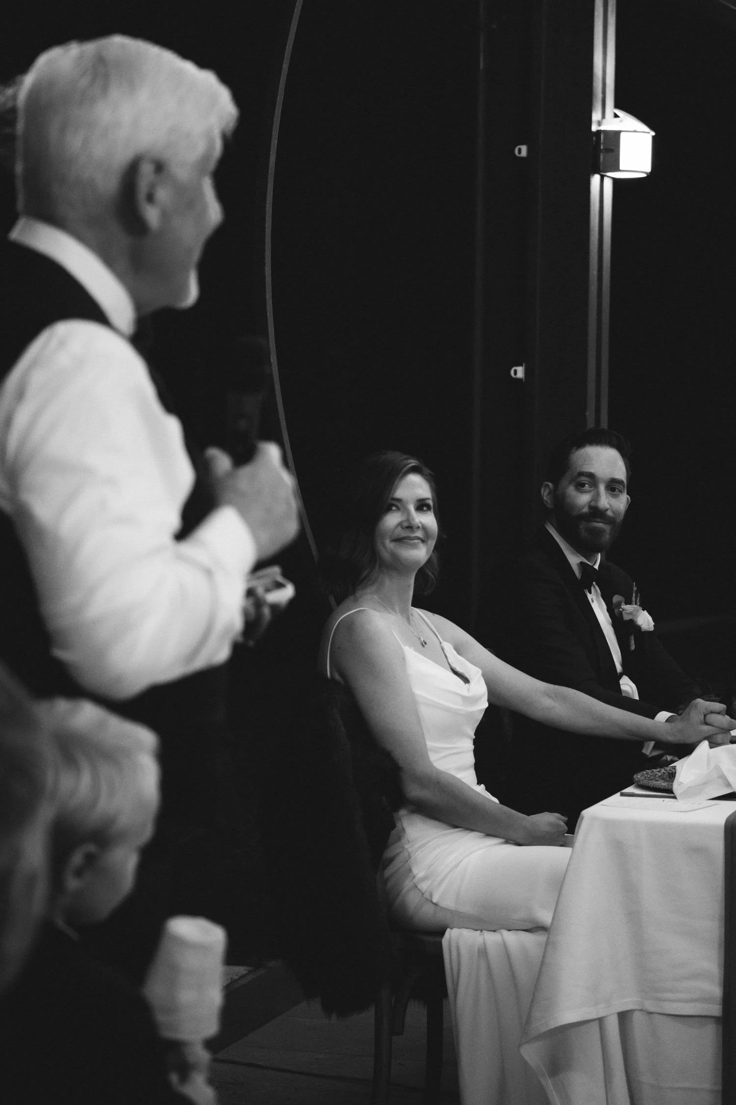 Black and white photo of bride and groom seated at reception table listening to a guest speech