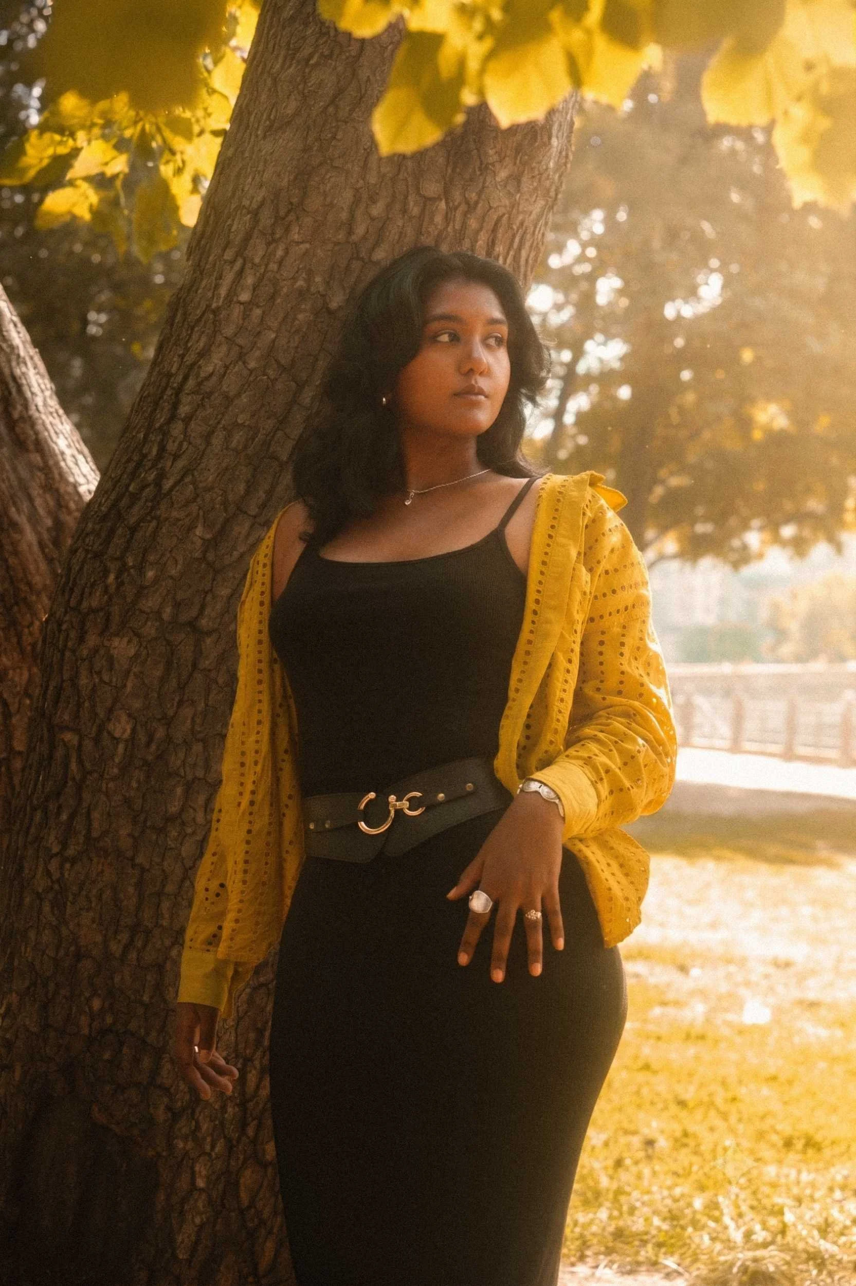 A portrait of a woman in a black dress and yellow shirt leaning against a tree during golden hour.