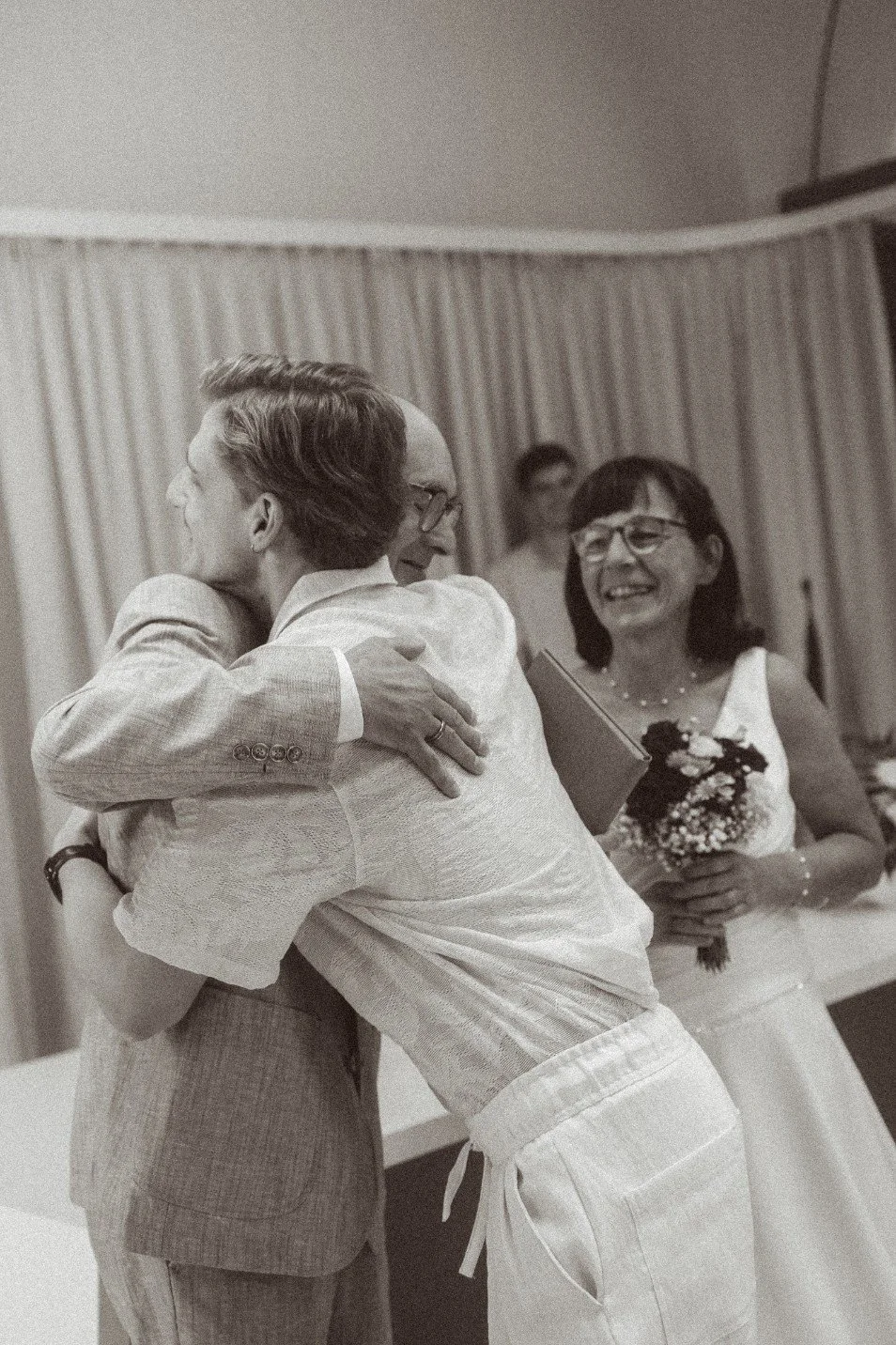 A black and white photo of a wedding guest hugging the groom while the smiling bride looks on indoors.