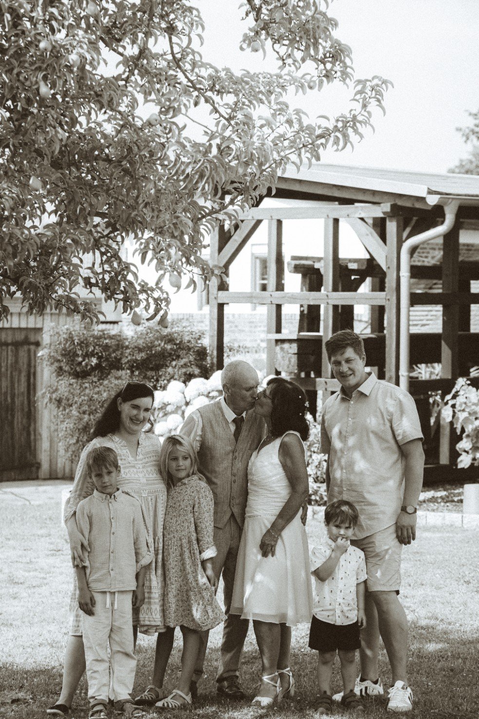 A black and white family portrait capturing a bride and groom kissing surrounded by family outdoors.