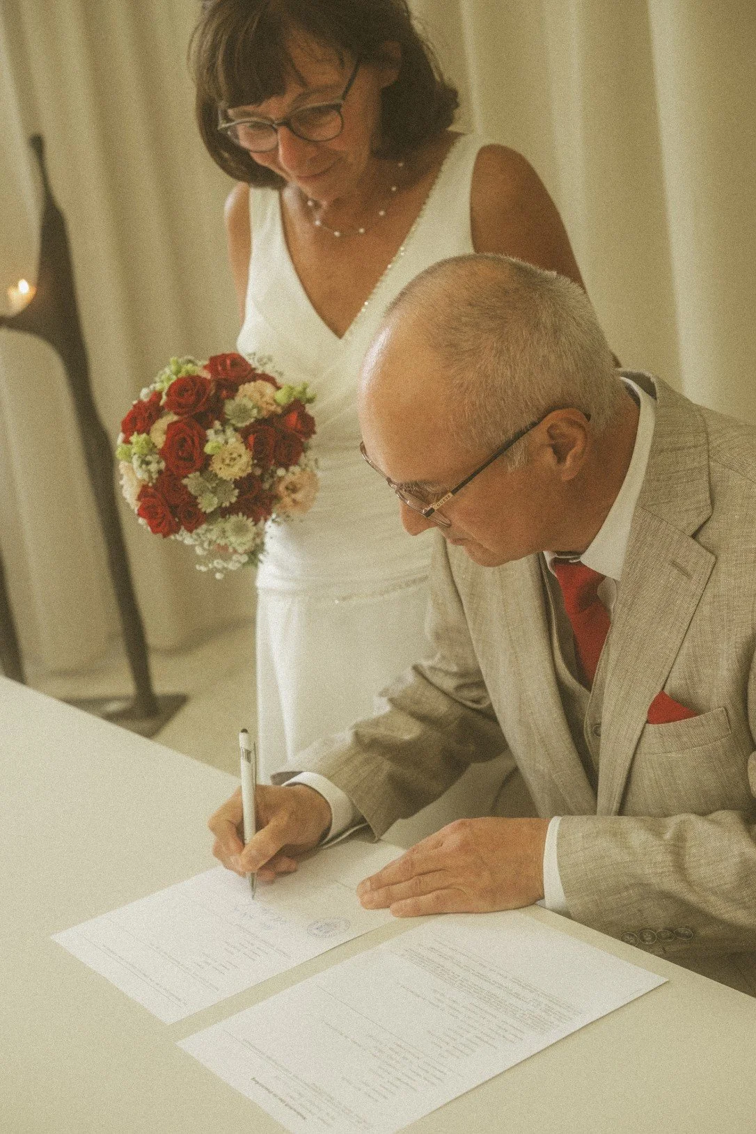 A groom signing a document at a table while the bride holding a bouquet stands behind him during an indoor wedding ceremony.