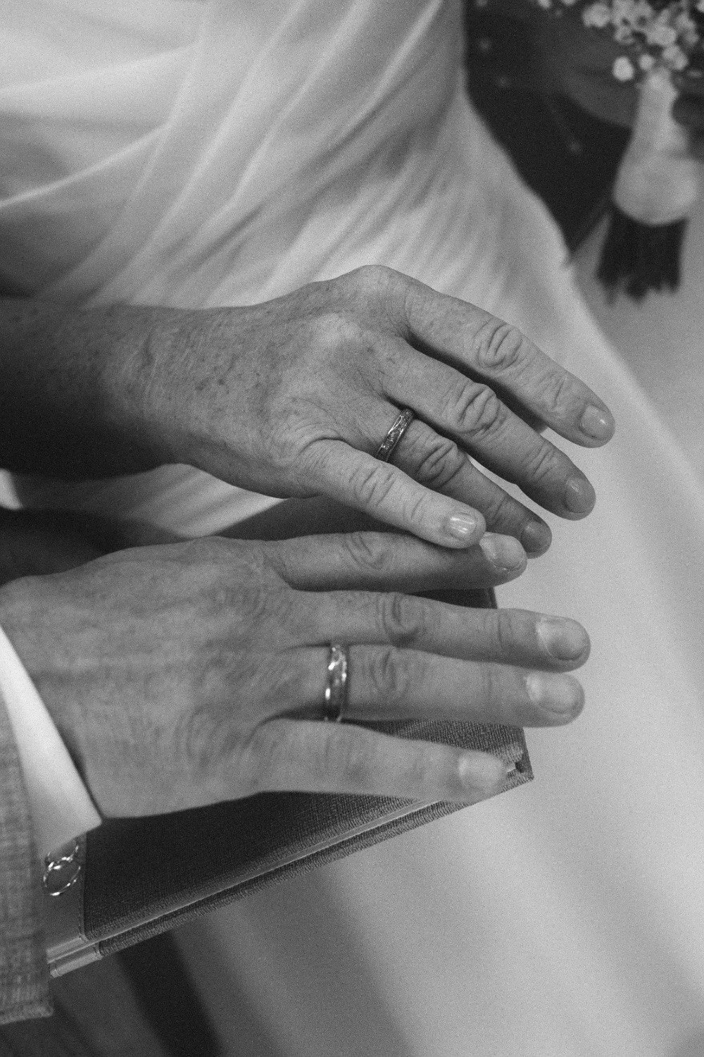 A close-up black and white indoor shot of the newlywed couple's hands resting together, proudly displaying their wedding bands.