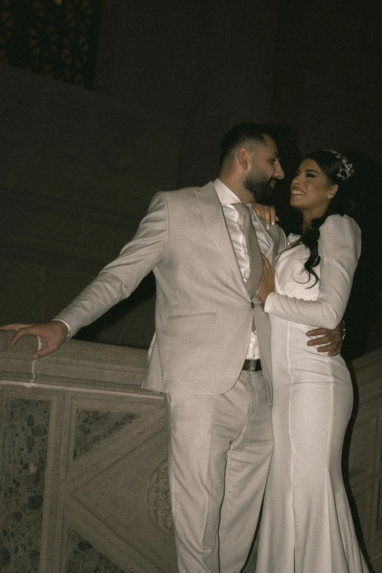 A couple in wedding attire embracing on a balcony at night, smiling at each other.