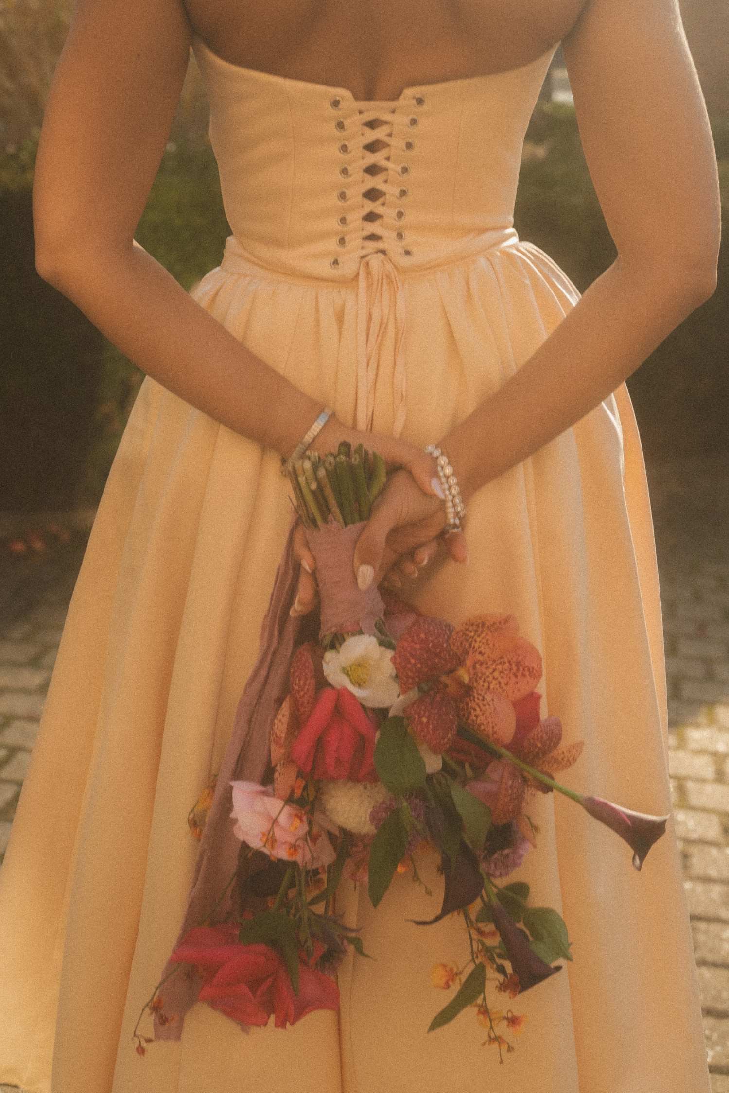 Close-up detail of a bride holding her bouquet behind her back, showing the laced wedding dress and floral arrangement.