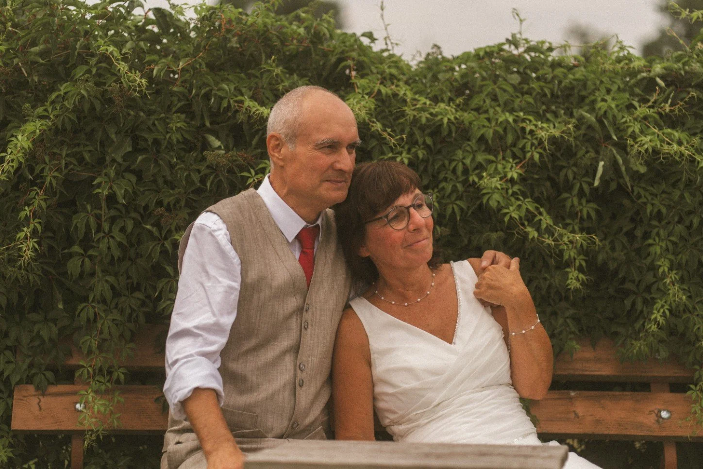 A newlywed couple sitting closely together on a wooden bench against a backdrop of lush green foliage.