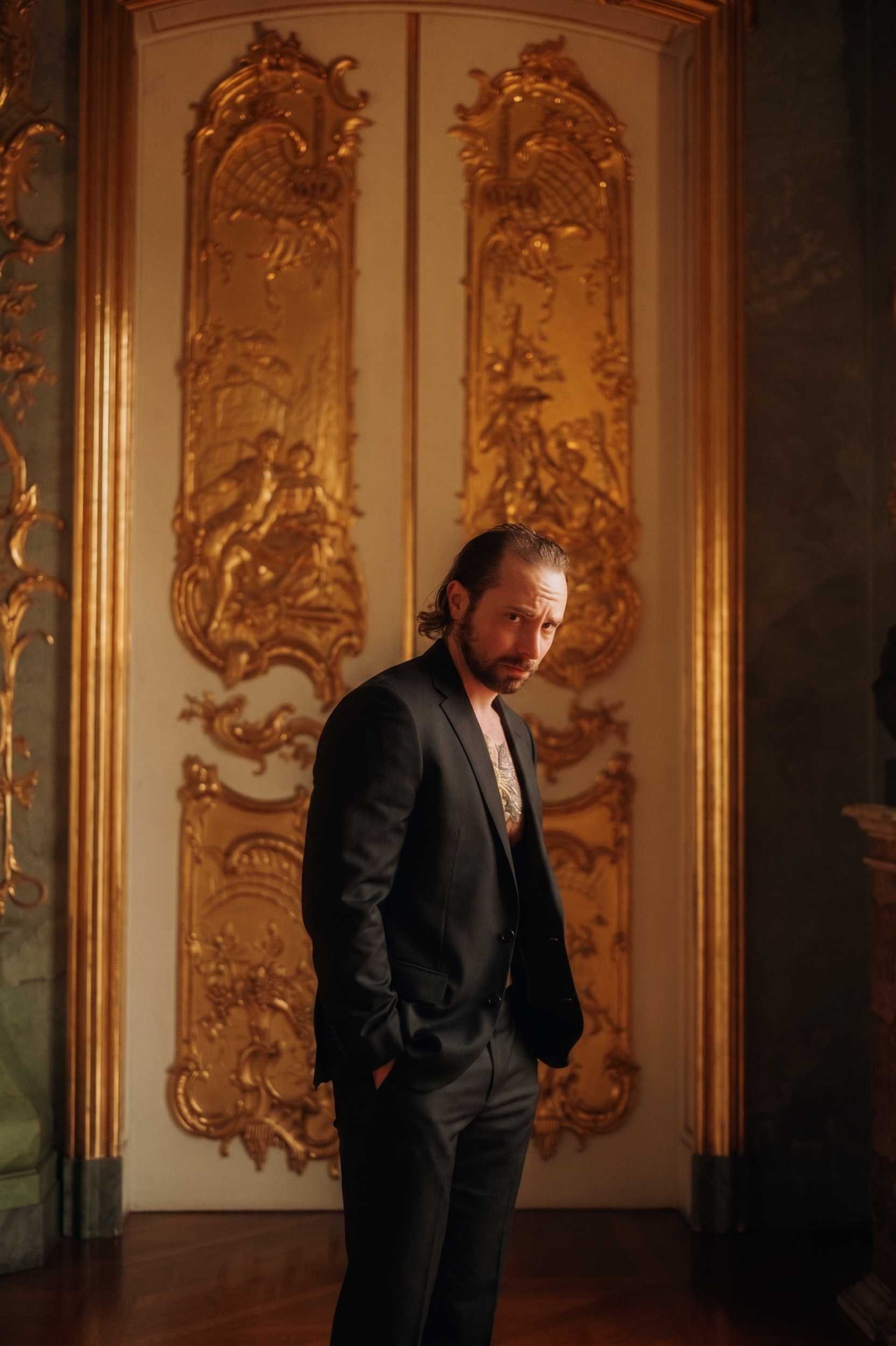 A man in a sharp black suit posing confidently in front of grand, ornate golden doors.