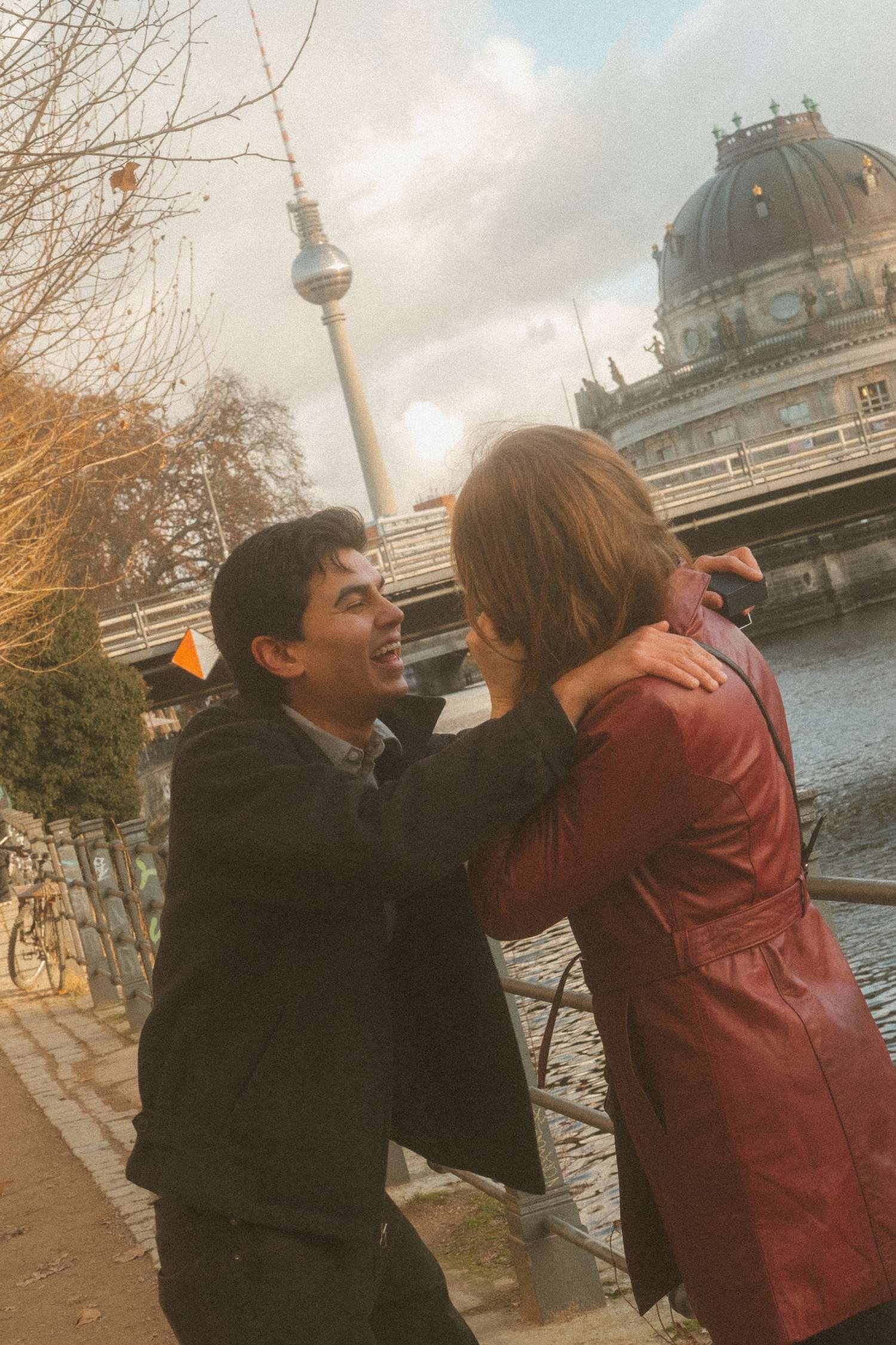 Laughing man embracing his partner by the Spree River with the Berlin TV Tower and Bode Museum in the background during their engagement photo session