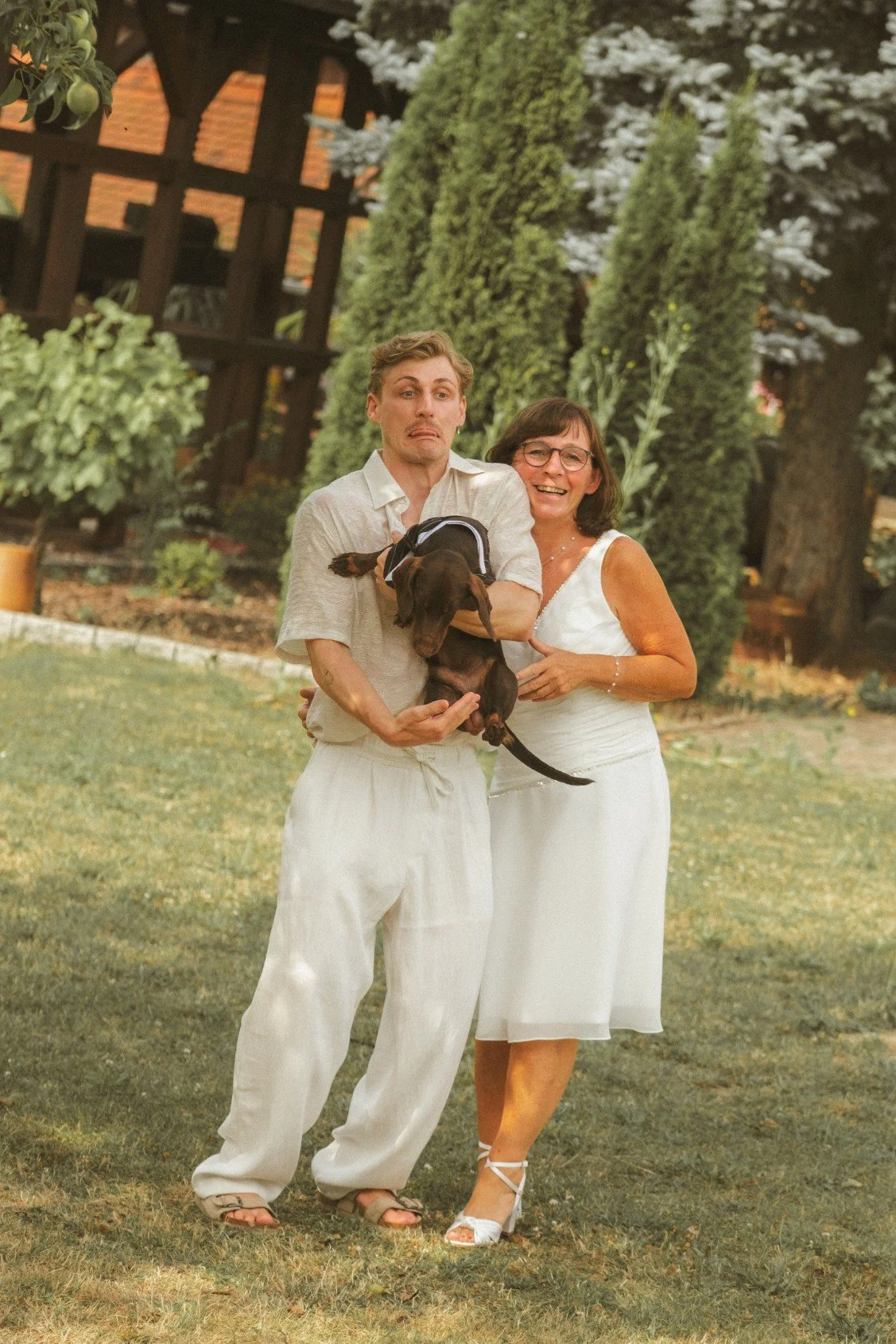 A medium shot of a young man making a funny face while holding a small dog, standing outdoors on grass next to the smiling bride in a white dress.