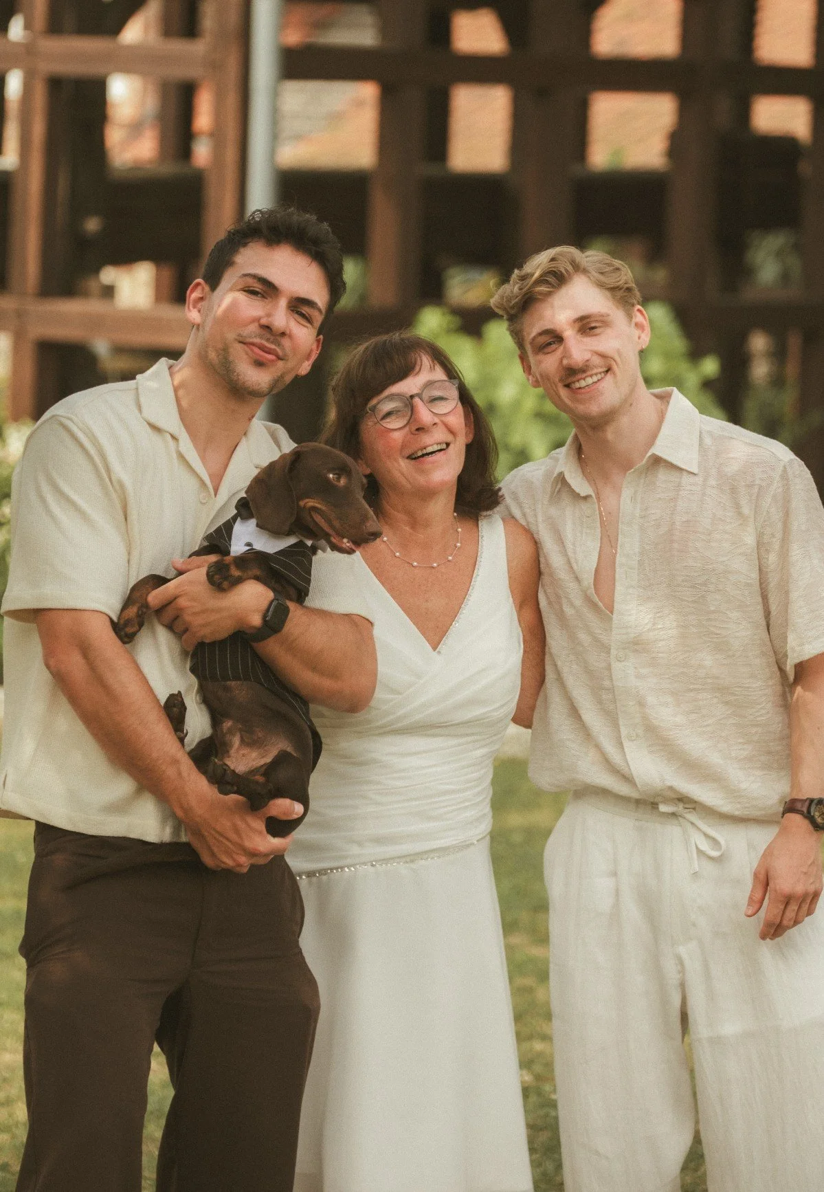 A medium outdoor portrait of two young men and the bride smiling together, with one man holding a small dog dressed in a tiny suit.