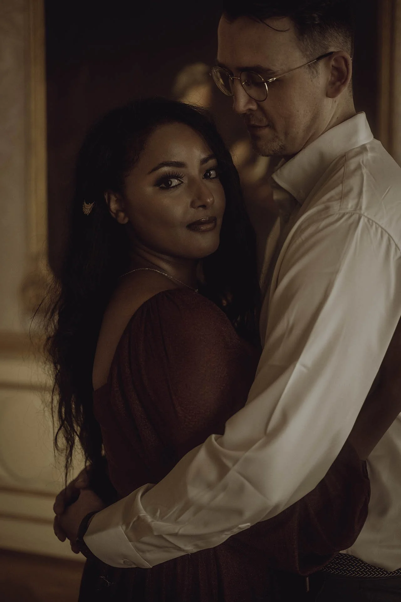 An intimate engagement portrait captured at Charlottenburg Palace in Berlin, showcasing a couple in a warm and romantic embrace. The bride-to-be radiates confidence in a burgundy gown, complemented by her fiancé’s elegant white shirt. This image high