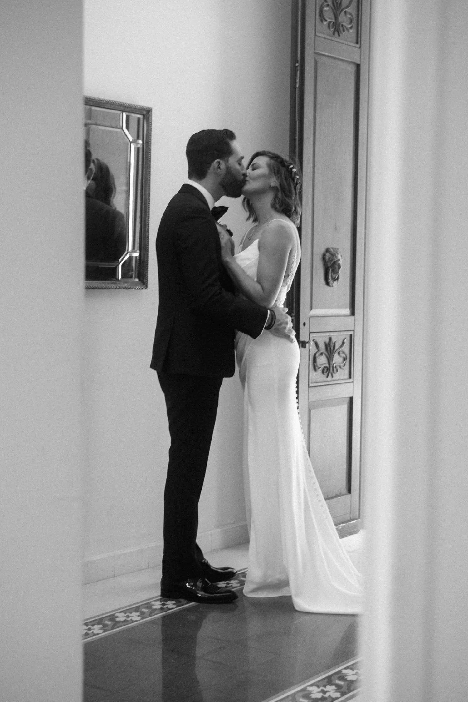Black and white wedding photo of a bride and groom sharing a quiet kiss inside an elegant interior hallway.