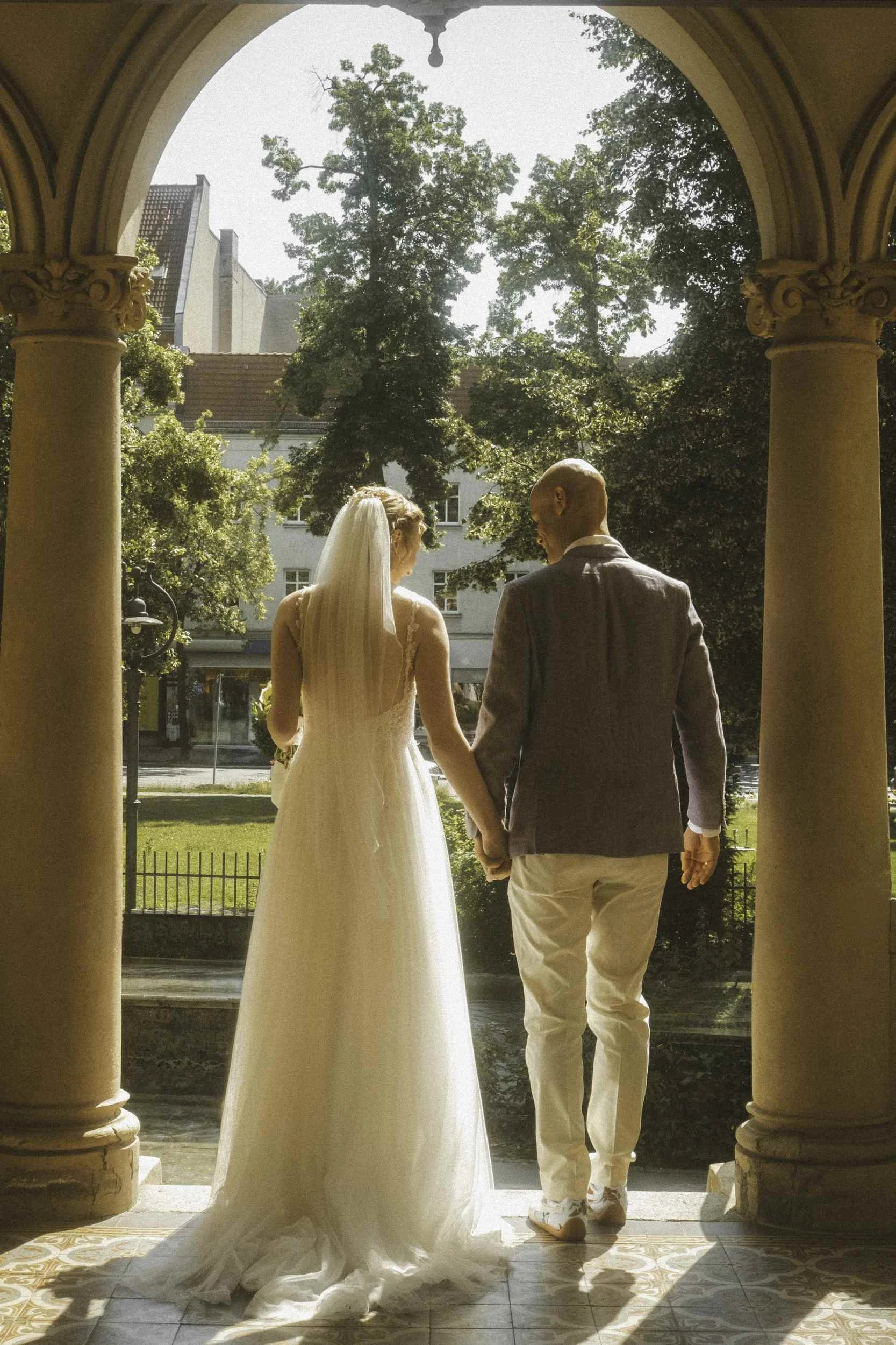 Newlyweds from behind hand in hand framed by ornate stone archways looking into Berlin Standesamt garden
