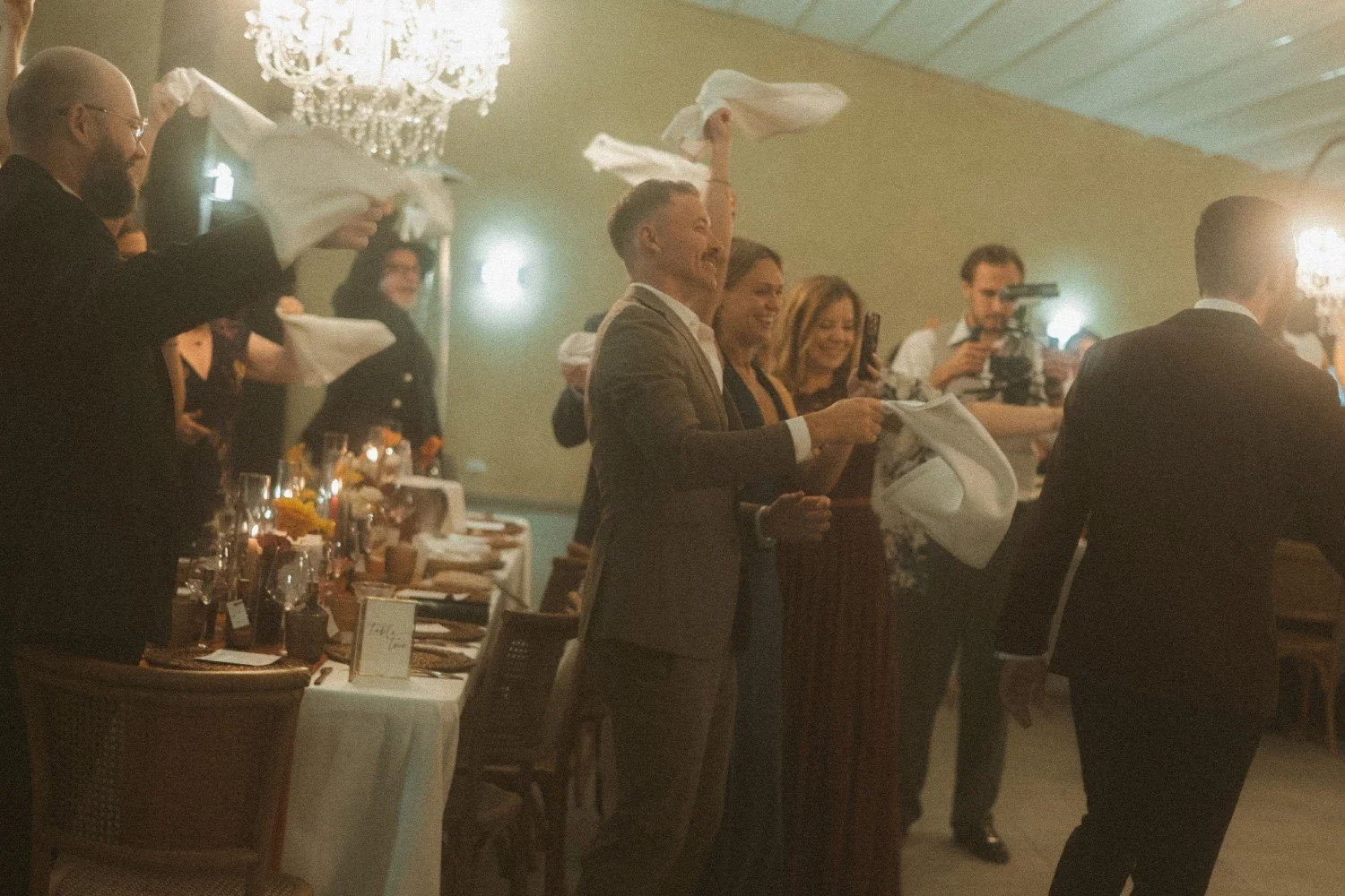 Wedding guests waving napkins and cheering during lively reception celebration indoors