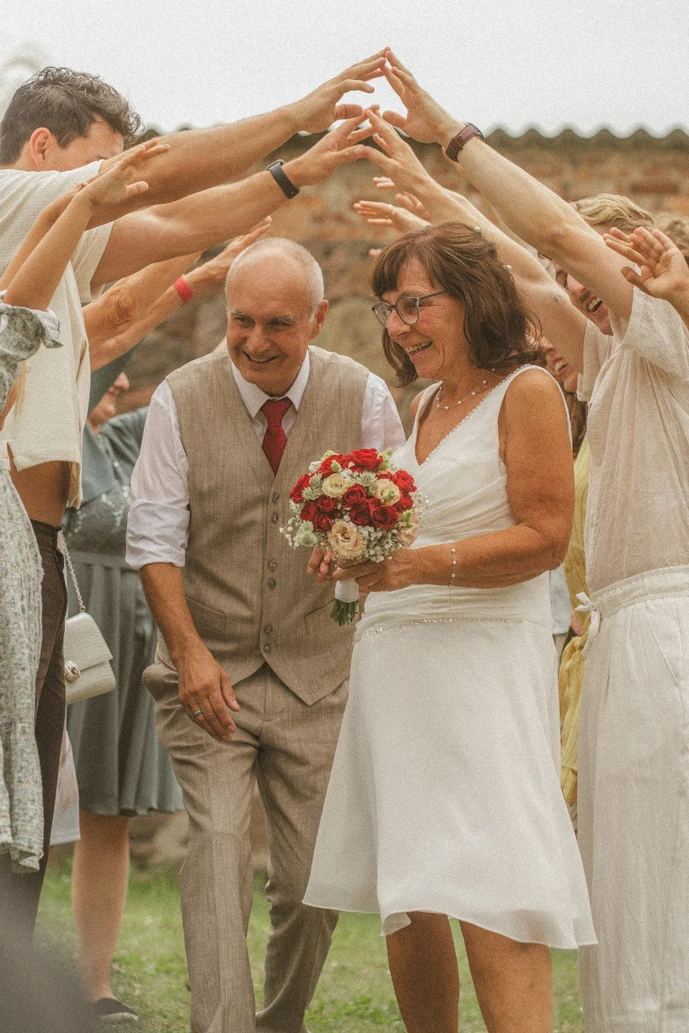 A medium outdoor shot of the smiling wedding couple walking underneath an arch formed by the raised arms and hands of their guests.