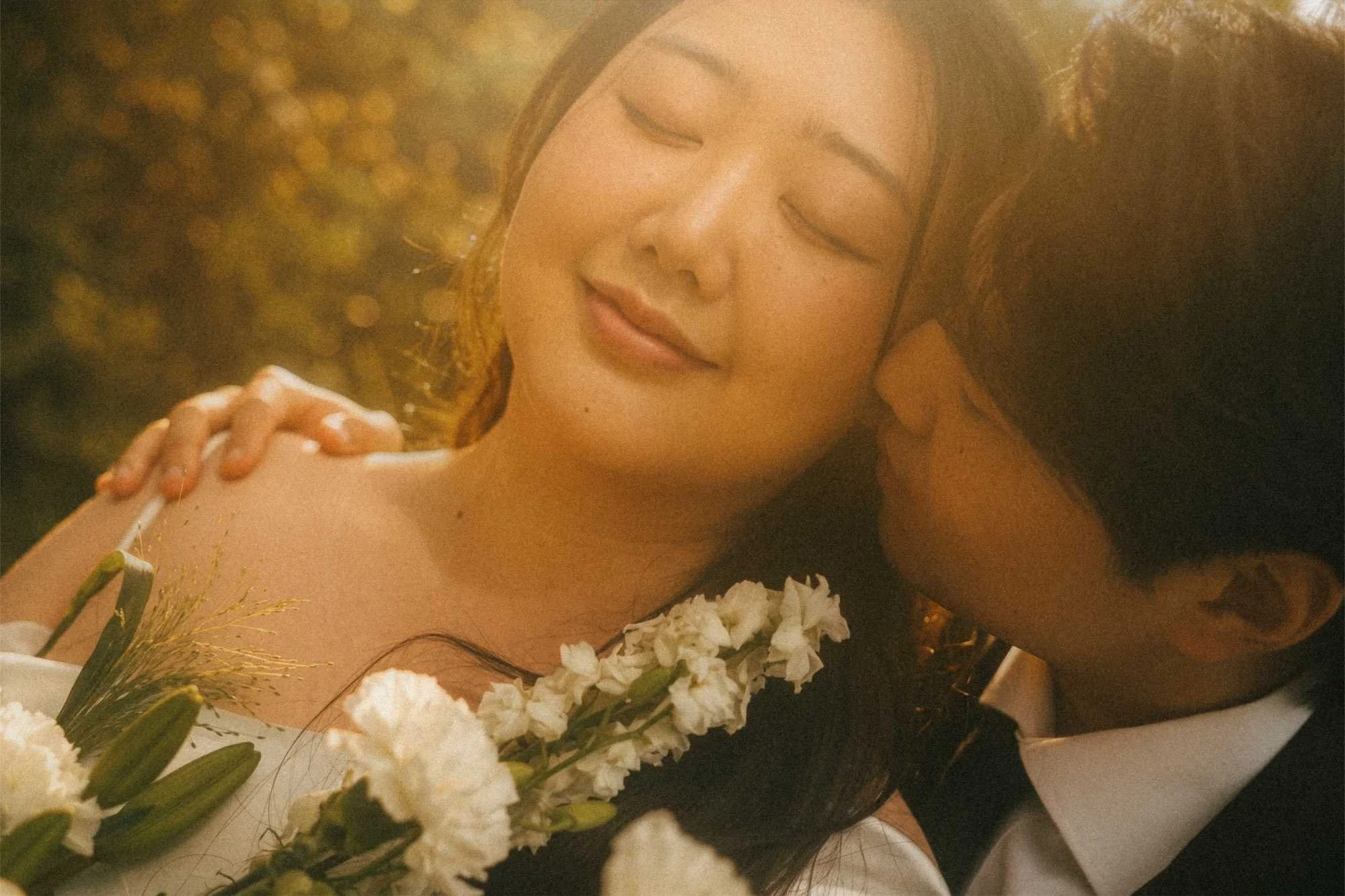 Close-up portrait of a groom kissing a smiling bride on the cheek as she holds a white floral wedding bouquet in golden hour sunlight in Potsdam.
