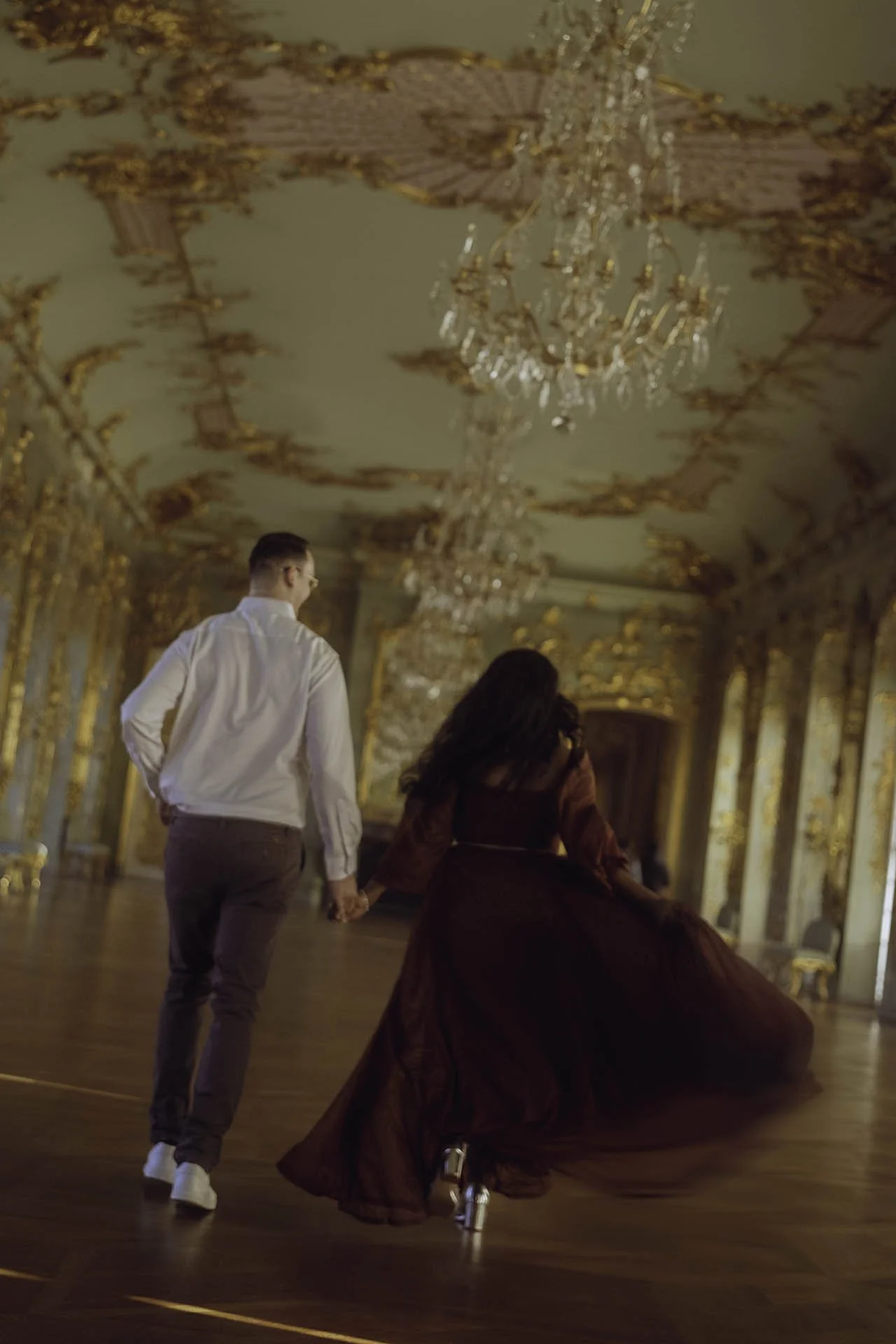 A stunning engagement photo of a couple walking hand in hand through the magnificent golden hall of Charlottenburg Palace in Berlin. The elegant maroon dress flowing behind the woman complements the grandeur of the room's ornate chandeliers and intri