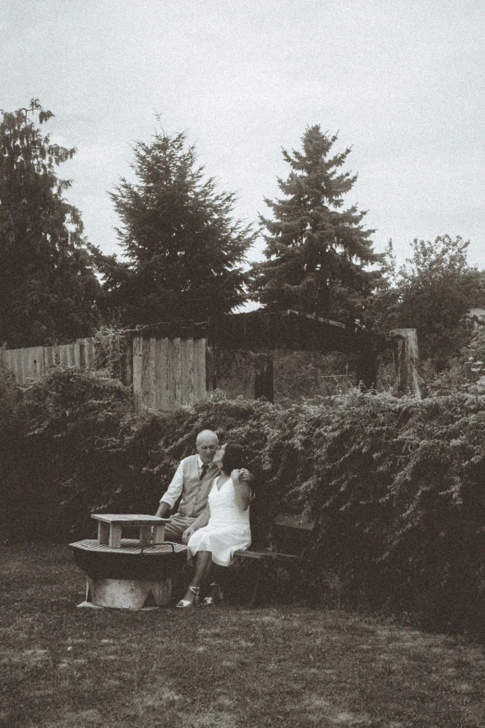 A wide black and white outdoor shot of the wedding couple sitting together on a bench next to a fire pit in a garden.