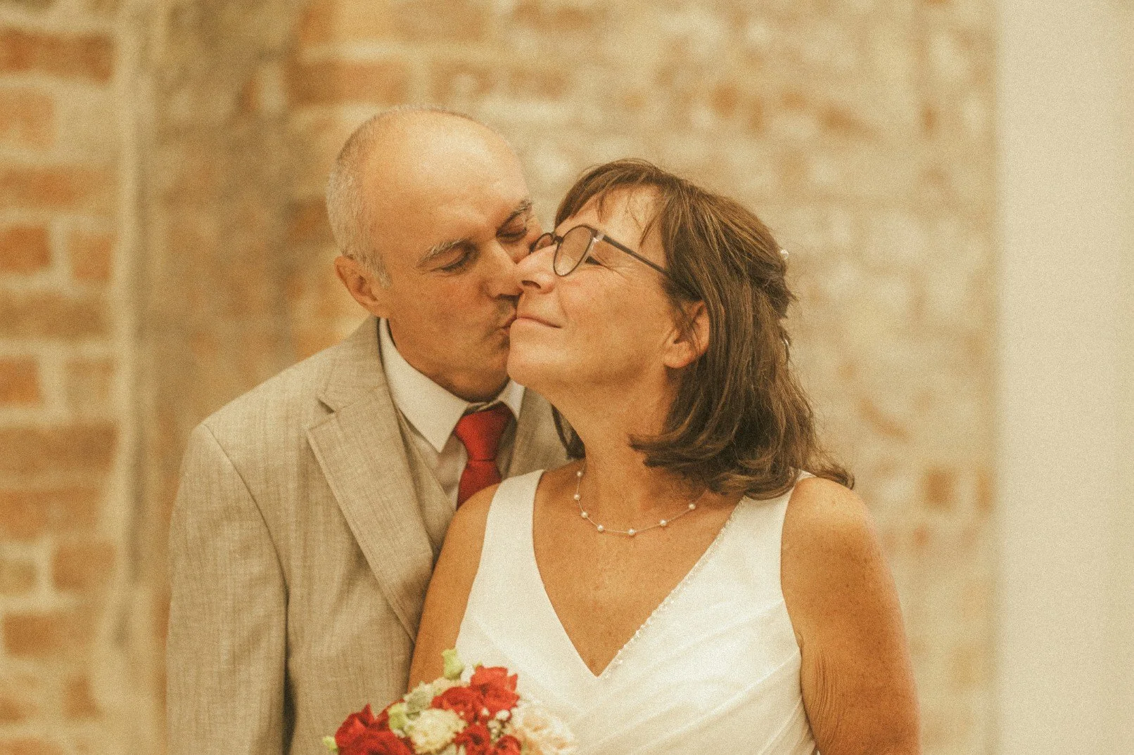 A close-up portrait of the groom gently kissing the smiling bride on the cheek against a brick background.
