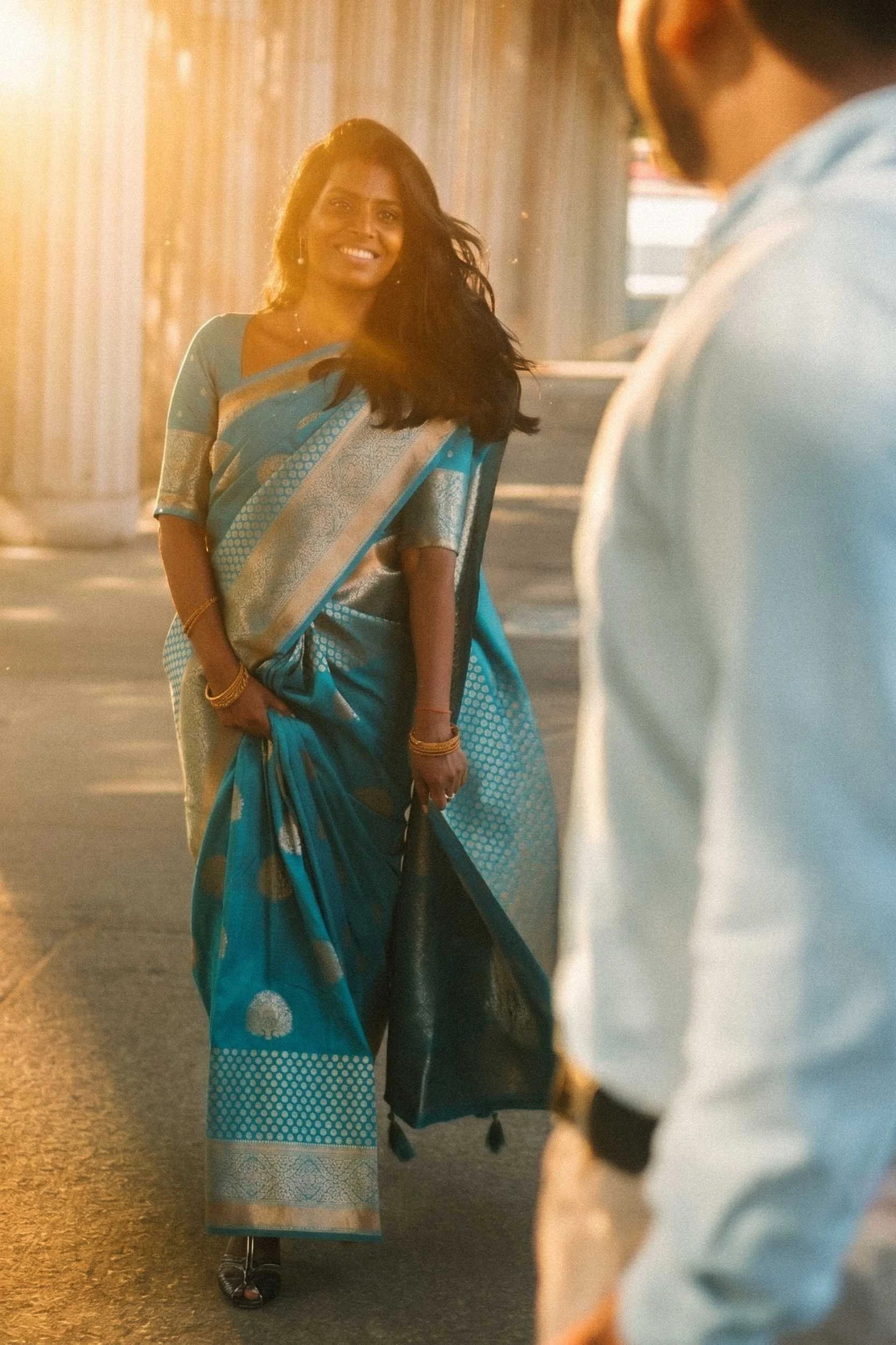 A woman wearing a traditional teal and gold silk sari smiles during golden hour, with a person in the foreground.