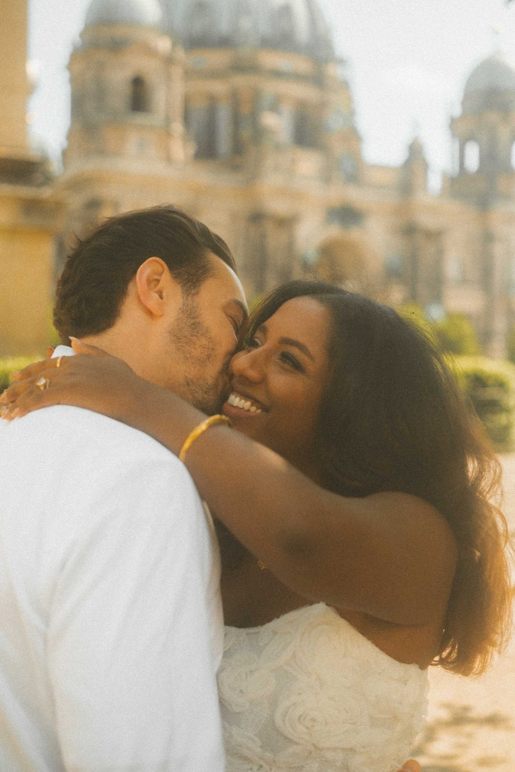 A romantic engagement portrait of a couple embracing in Berlin, with the groom kissing the bride on the cheek while she smiles, set against the historic cathedral.