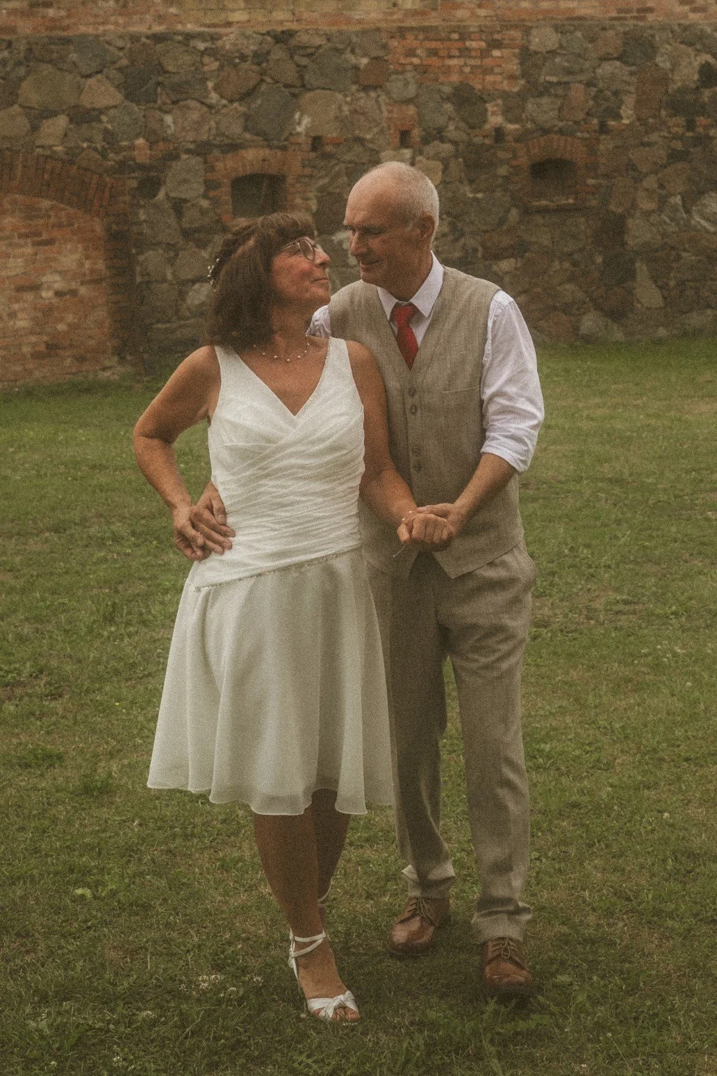 A bride and groom smiling at each other while walking together on the grass in front of an old stone wall.