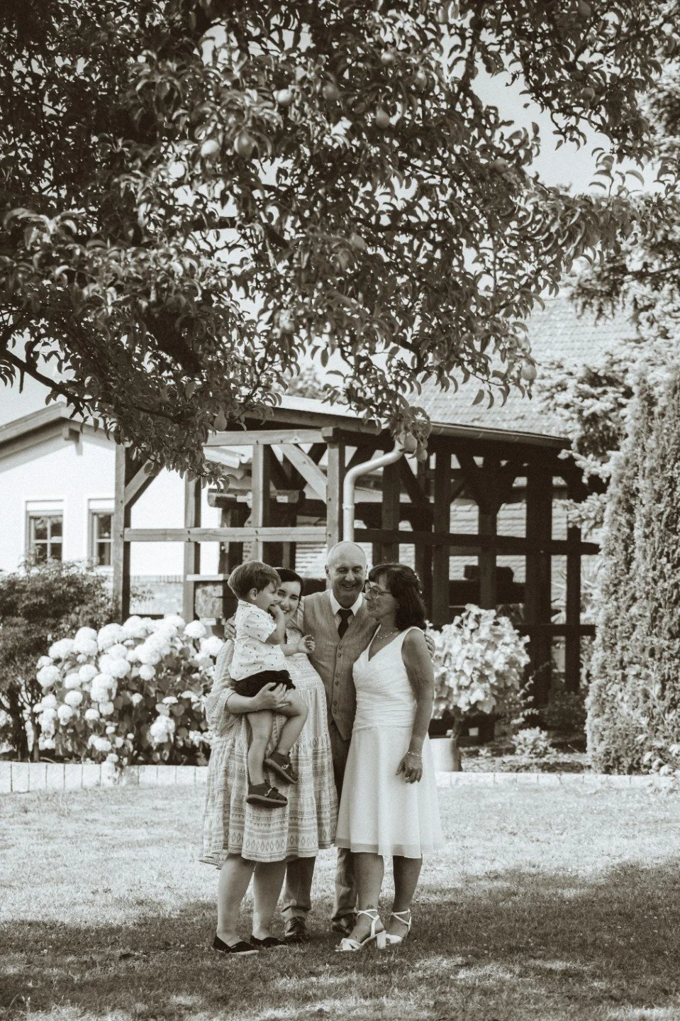 A medium black and white group portrait of the wedding couple posing outdoors with a woman holding a young child near a wooden structure.