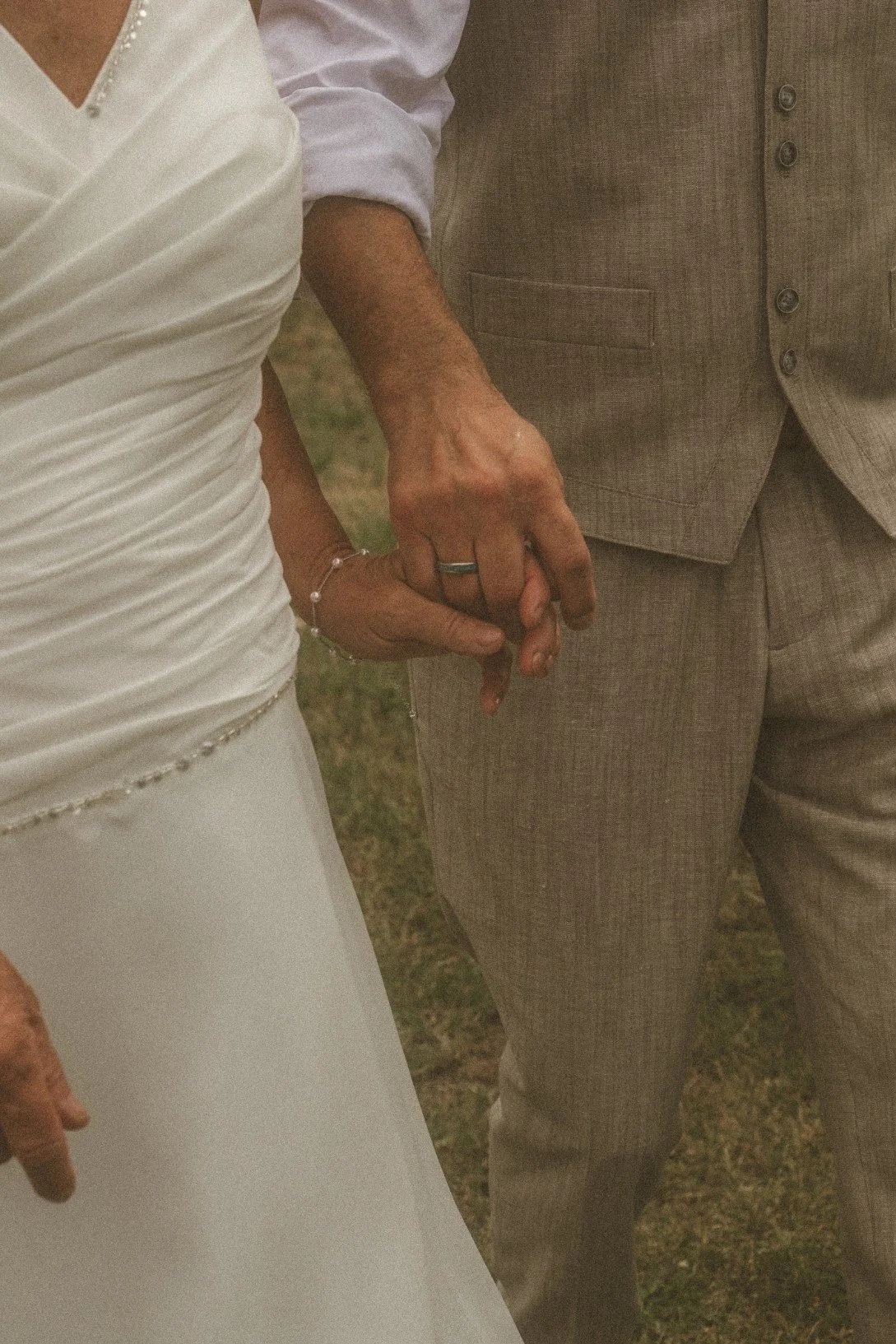 A close-up shot of a bride and groom holding hands, highlighting their wedding bands and attire outdoors.