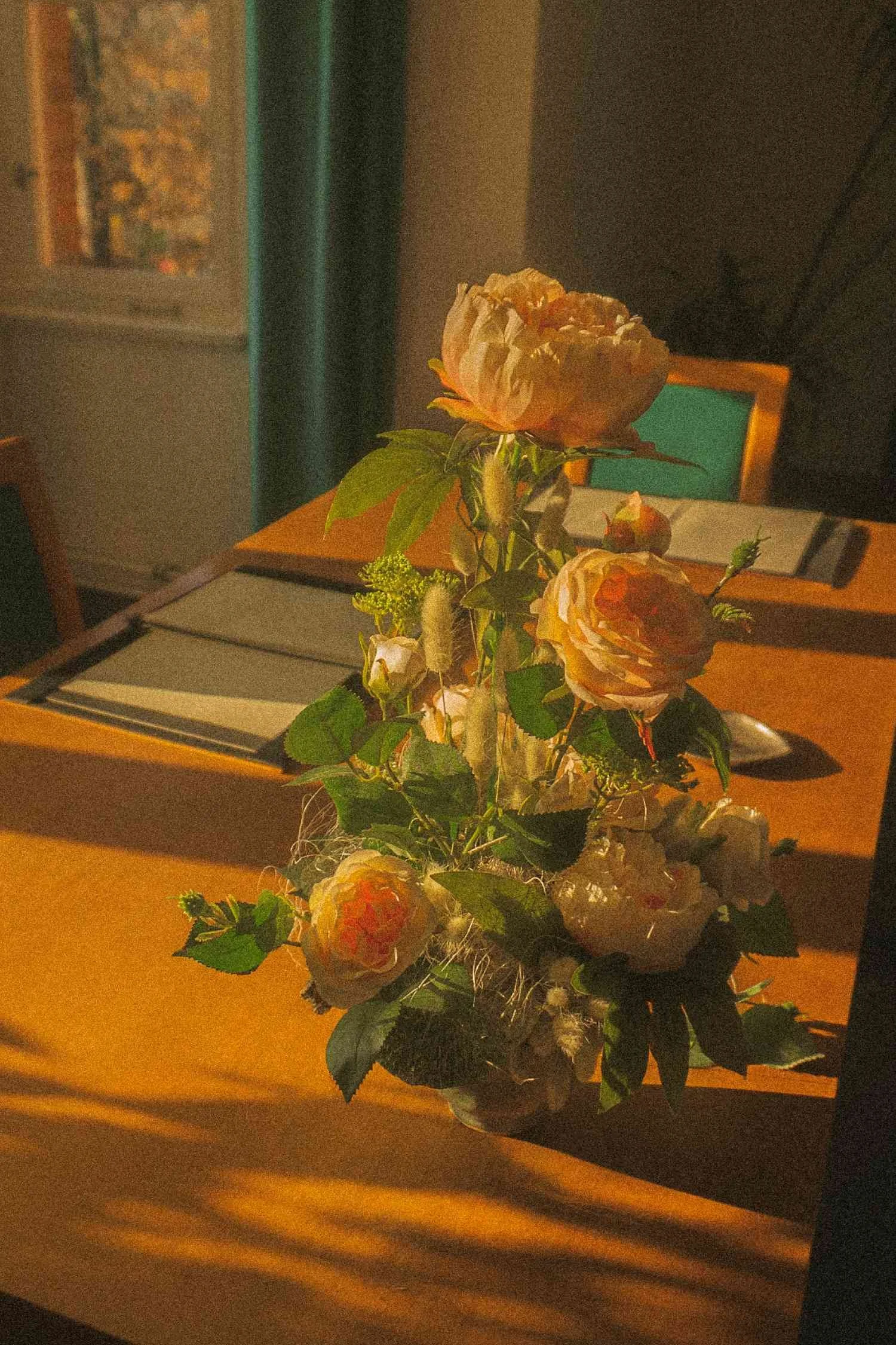 Still life of a floral arrangement resting on a wooden table, lit by warm window light with chairs softly out of focus behind.