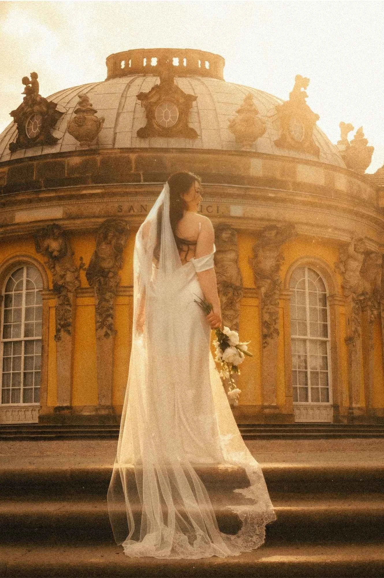 Rear view of a bride in a long flowing veil and white gown holding a bouquet, standing on stone steps in front of the historic Sanssouci Palace dome in Potsdam.