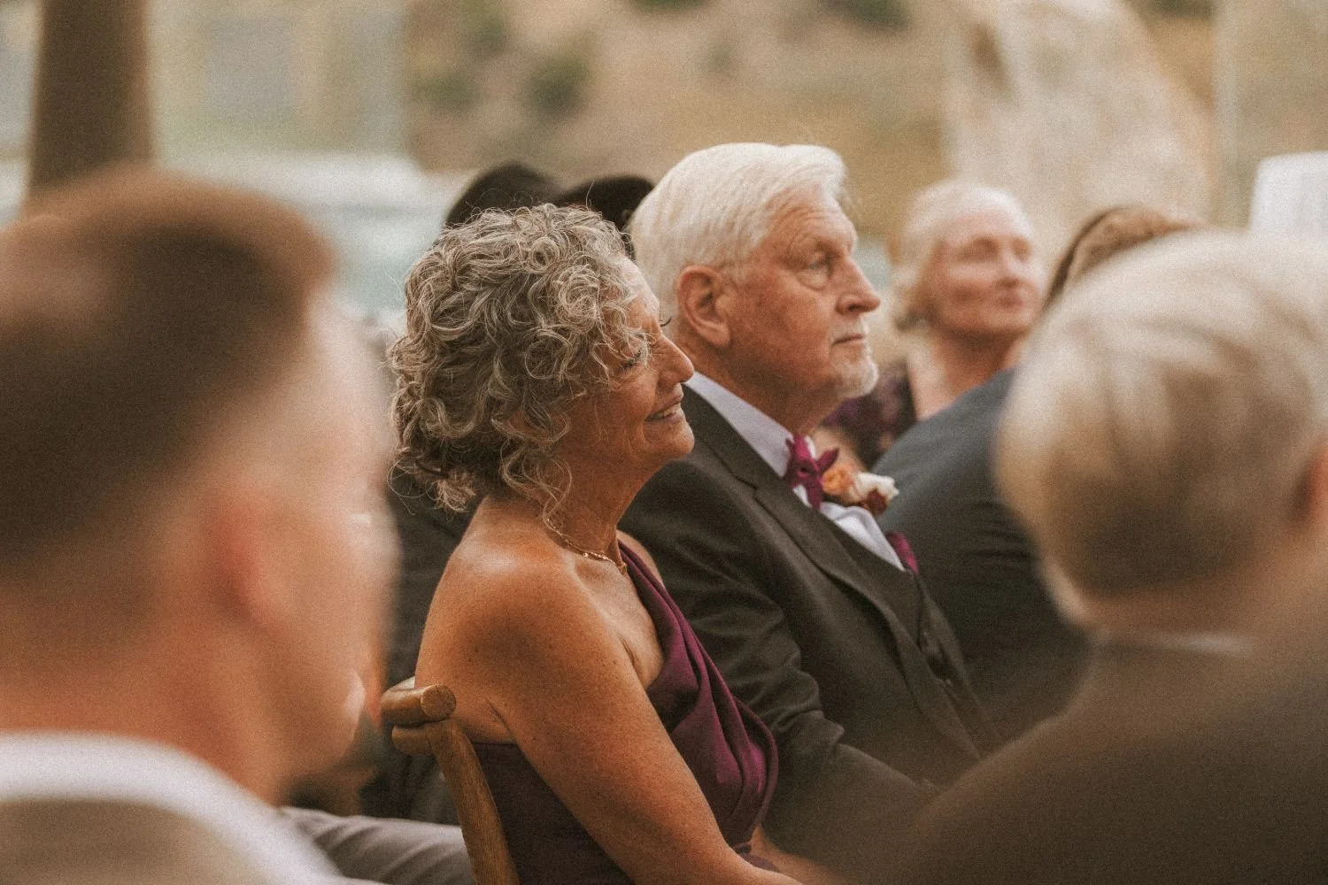 Elderly couple seated together during a wedding ceremony, sharing a quiet emotional moment among guests.