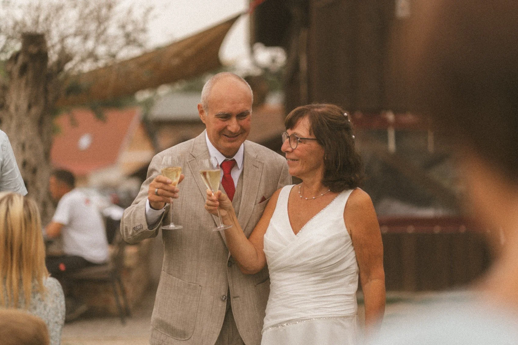 A newlywed couple smiling and raising glasses for a toast during an outdoor wedding reception.