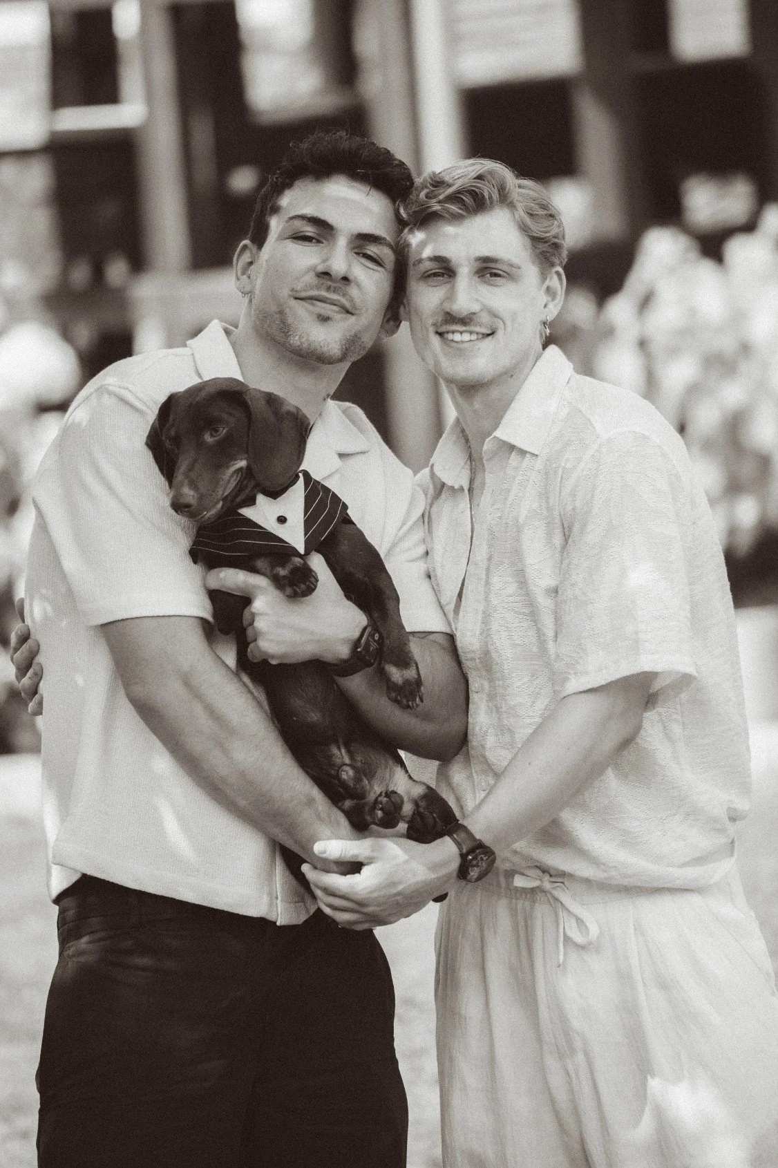 A medium black and white outdoor portrait of two smiling young men holding a small dog dressed in a tiny suit.