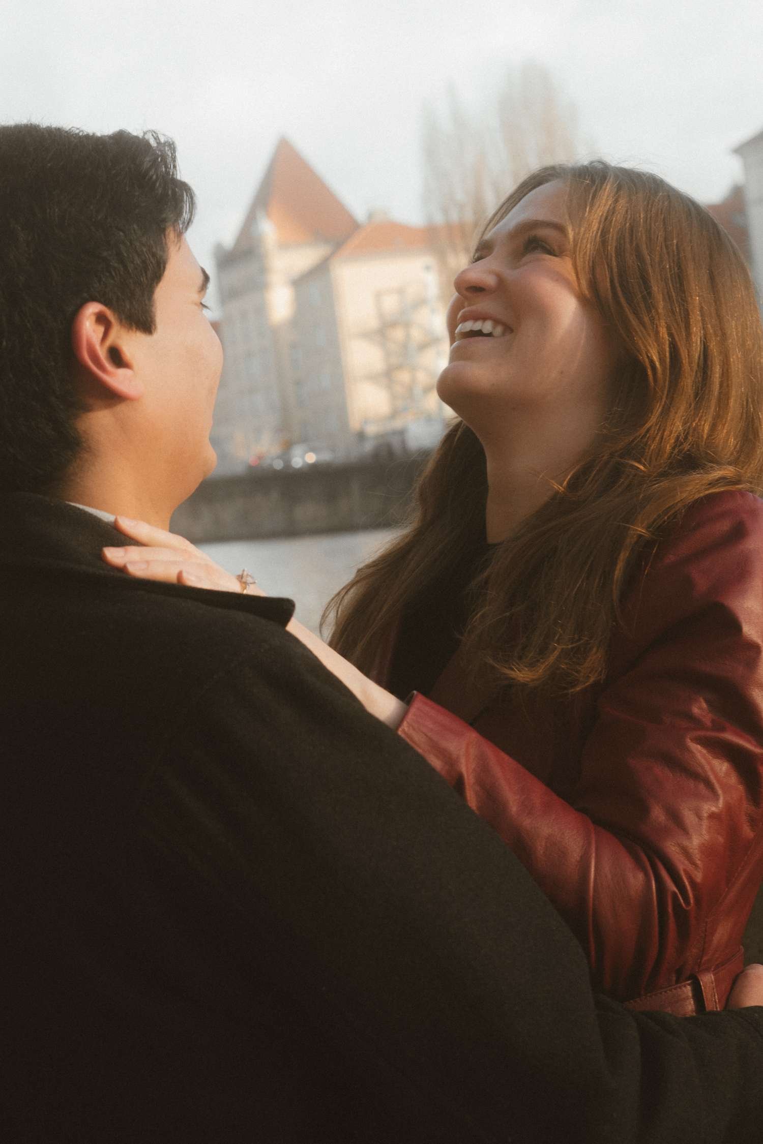 Laughing woman looking up at her partner with historic Berlin buildings and a river visible behind them during their engagement photo session