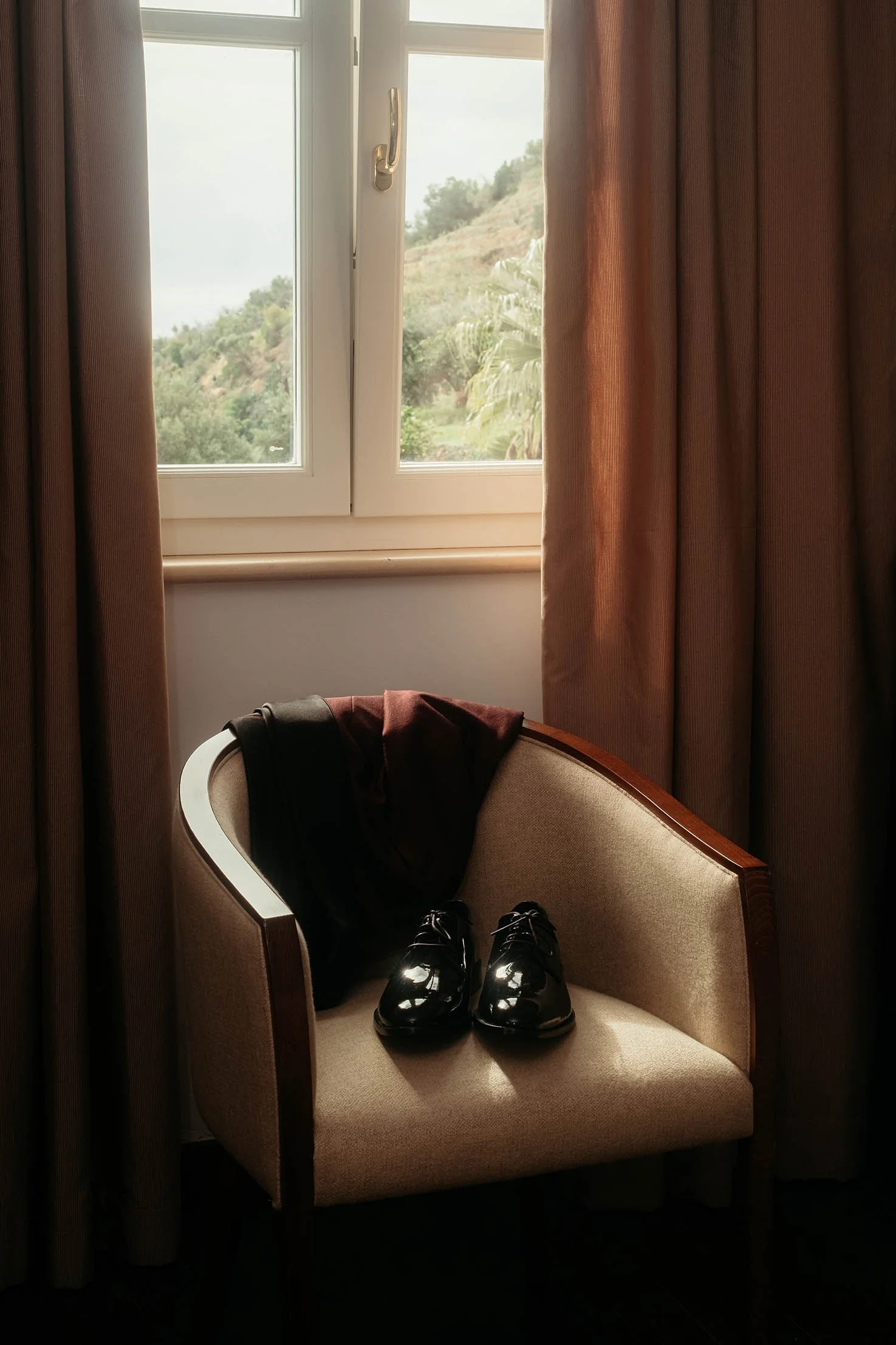 Groom’s polished shoes and suit jacket placed on a chair by the window during wedding preparations in Sicily.