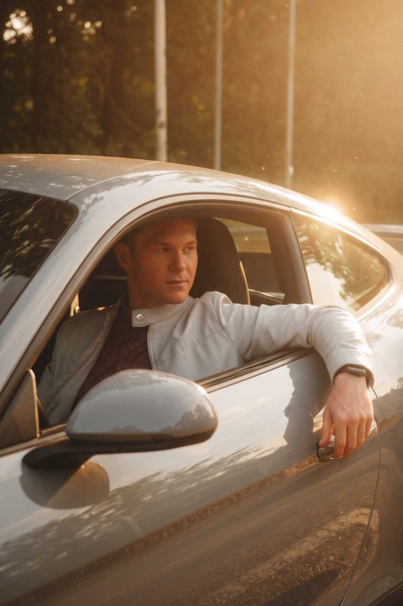 A man in a light jacket driving a sports car, looking out the window during golden hour.