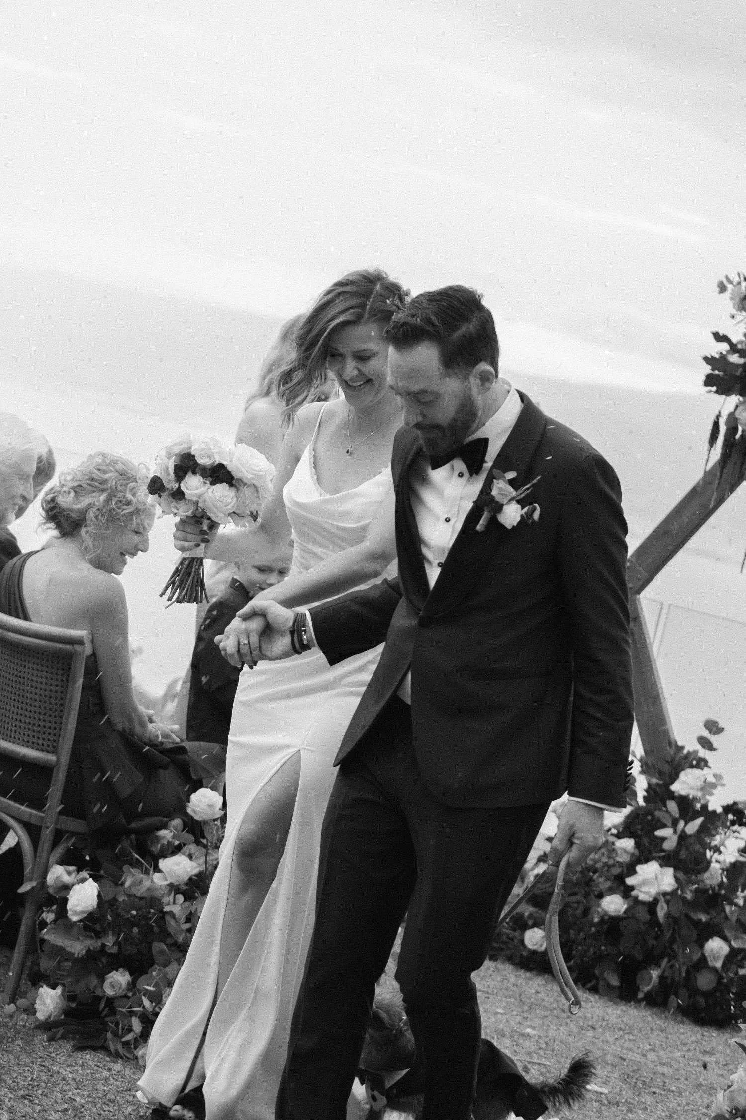Bride and groom holding hands while walking down the aisle after an outdoor wedding ceremony in black and white