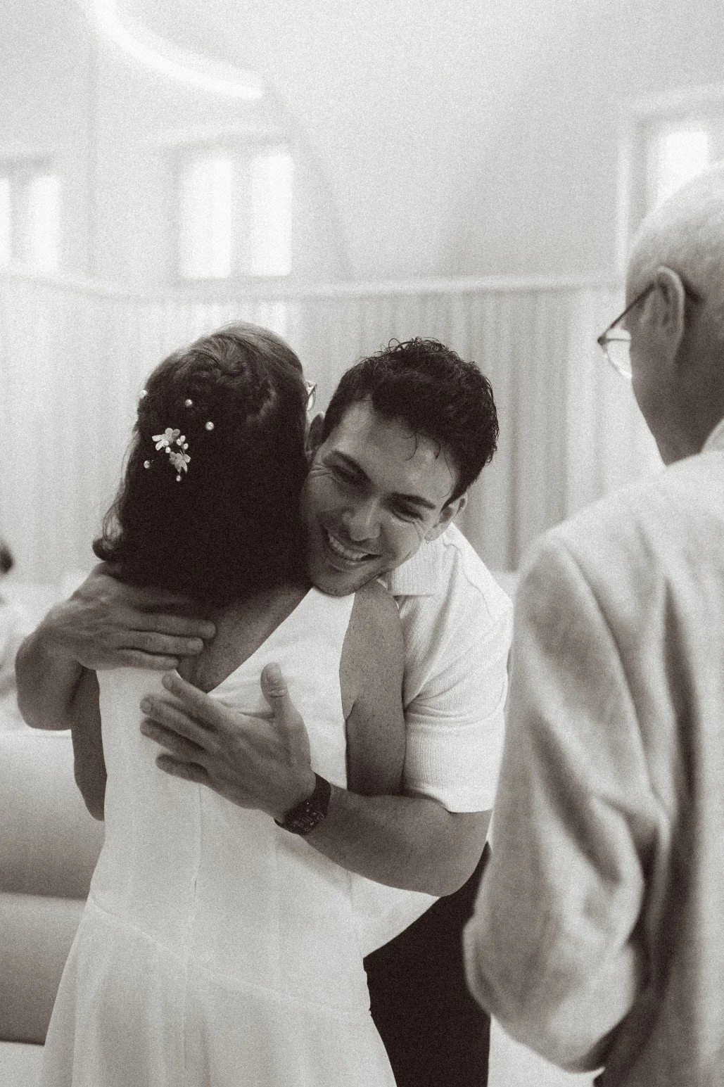 A black and white photo of a smiling wedding guest giving the bride a warm hug indoors.